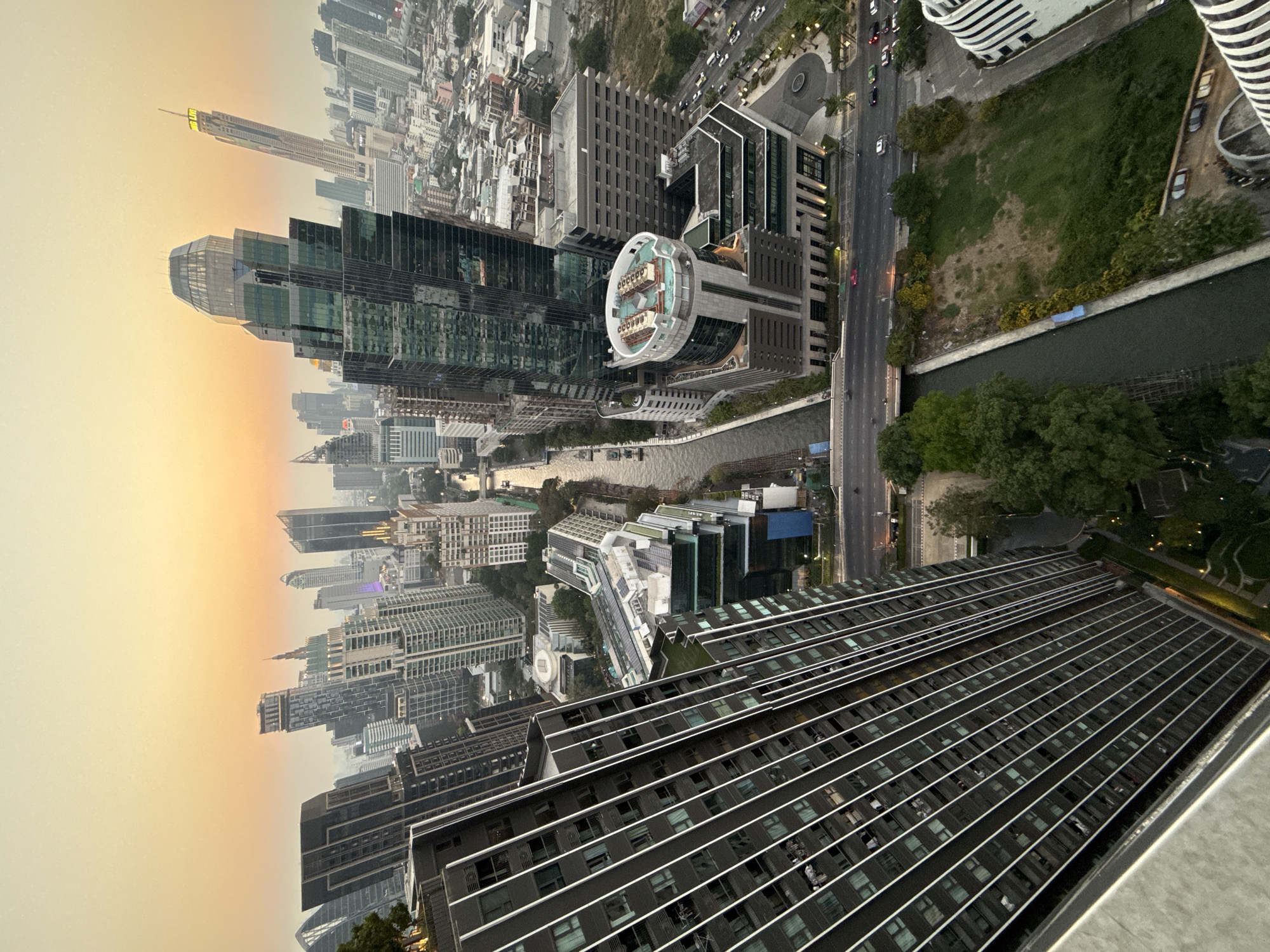 The image shows an aerial view of Bangkok, Thailand. Skyscrapers and other buildings dominate the urban landscape, with a river running through the city. A street with vehicular traffic is visible beside the river. The foreground features the top of a building with a linear pattern of windows. The sky is hazy, possibly due to pollution or weather conditions.