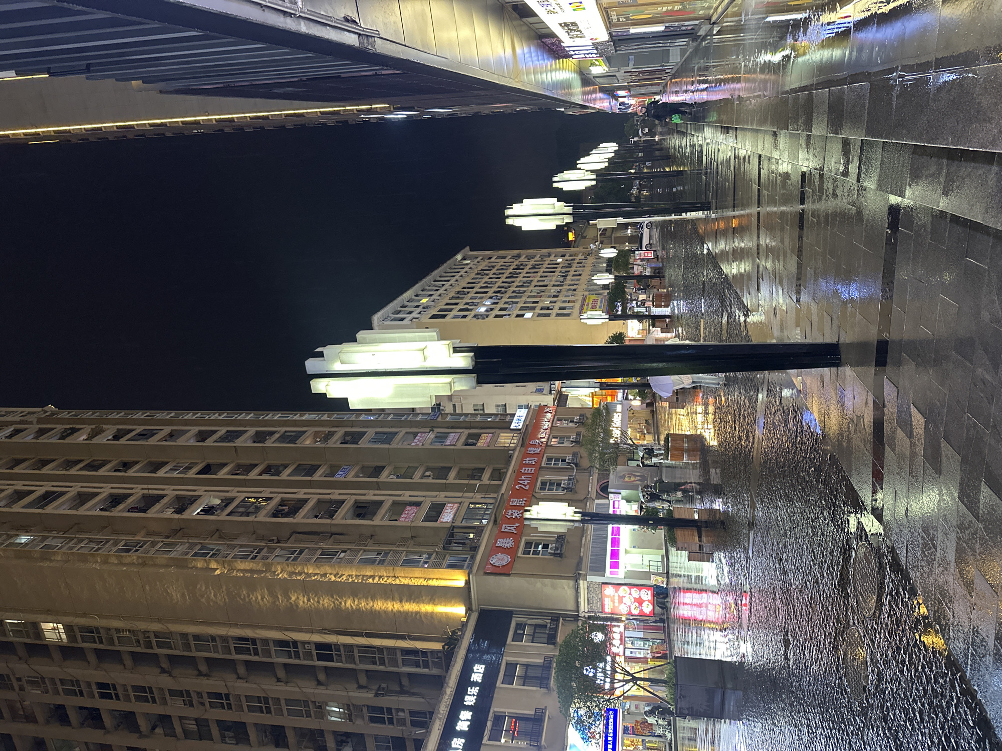 This media file captures an urban street scene at night in Chongqing High-tech Zone, China, following recent rainfall. The setting is an illuminated pedestrian walkway bordered by tall buildings, creating a reflective and atmospheric environment.
In the foreground and midground, a wide, dark, and wet pavement stretches into the distance on the right, reflecting the bright artificial lights from surrounding structures and streetlamps. The reflections create elongated streaks of white, blue, and orange on the glossy surface, highlighting the texture of the paving stones. A series of modern, stacked cube-shaped streetlights line the right side of the walkway, emitting a cool white glow. Faint, blurred figures of people and possibly vehicles can be discerned further down the street.
To the left, tall, multi-story buildings dominate the frame. The one closest to the viewer is a light-colored building with numerous windows, some illuminated, suggesting residential or commercial spaces. Above this, a dark, metallic structure, possibly an elevated walkway or bridge, spans horizontally across the upper part of the image, with a strip of warm yellow light running along its underside.
Further along the street, another building features prominent signage. A long, red banner is visible, displaying Chinese text that reads "暴风袋鼠 24h自助健康" (Storm Kangaroo 24h self-service health) and "房 简餐娱乐 酒店" (Rooms, simple meals, entertainment, hotel). Other signs, including one with "年轻人更高光" (Young people are more radiant) and faint occurrences of "AME" and "00," are also present on the building facades.
The overall scene is vibrant with artificial light against a dark night sky. The wet ground is a notable detail, indicating recent rain and contributing significantly to the visual aesthetic through its reflective qualities. The combination of modern architecture, urban lighting, and a glistening wet surface lends the image a calm and contemporary city ambiance.