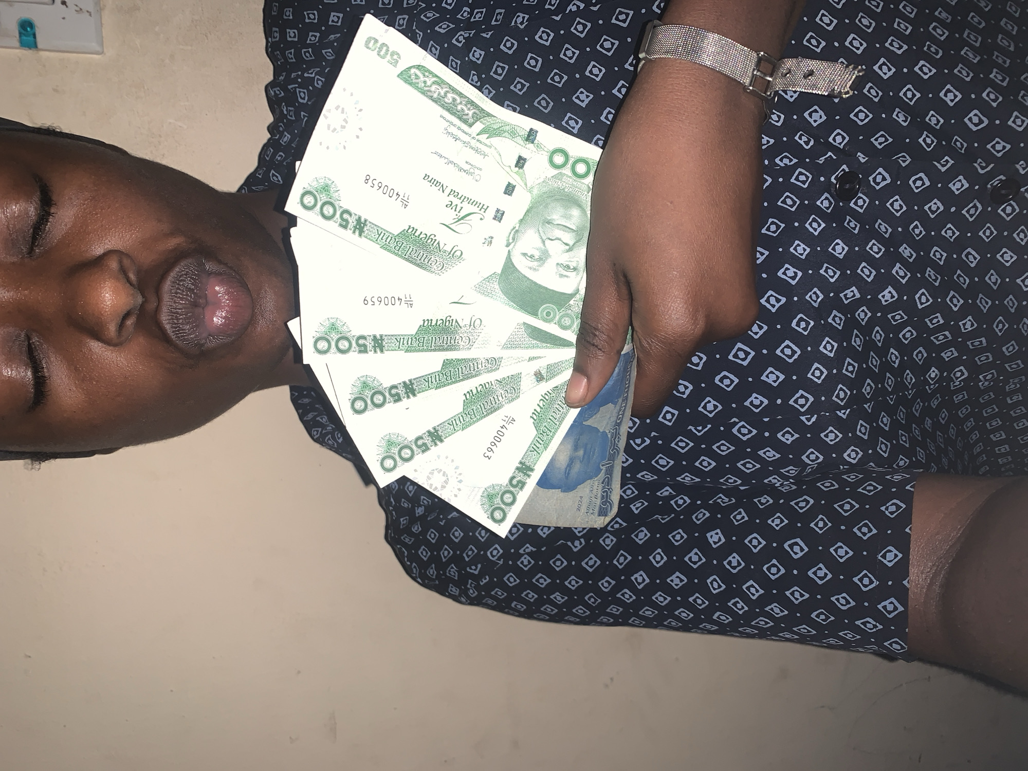 A person with closed eyes and pursed lips holds a fan of Nigerian 500 Naira notes in their hand. The person is wearing a dark blue shirt with a light blue geometric pattern and a silver watch. The background is a light-colored wall. There is no explicit indication of the location being in the city of New Stadium, Nigeria.