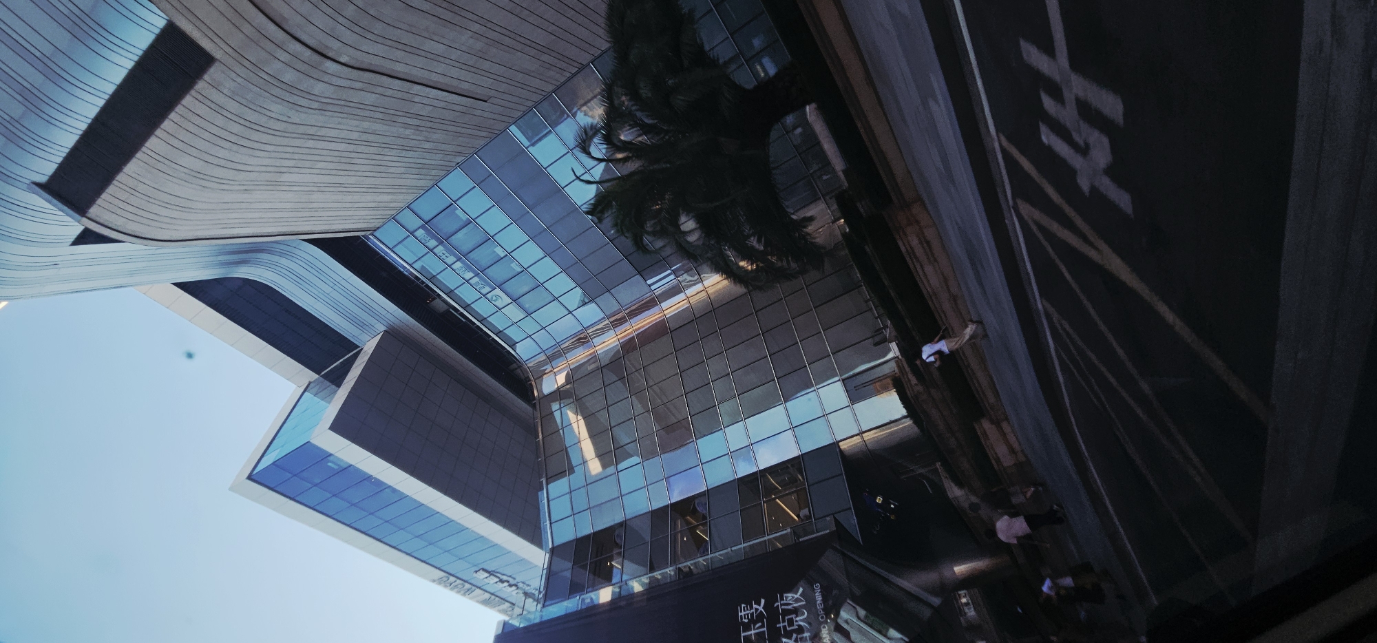 The image shows a low-angle, tilted view of a modern, multi-story building with a facade composed of glass and curved, textured concrete panels. The building's architecture is characterized by sharp angles and flowing lines, with reflective blue glass windows that mirror the sky and surrounding environment. In the foreground, a street with painted lines is visible, and several people are walking on the sidewalk. A palm tree with dark foliage is positioned to the right of the building. A black banner with Chinese characters and the words "GRAND OPENING" is affixed to the left side of the building. The sky is clear and blue. The scene appears to be in a commercial or urban area.