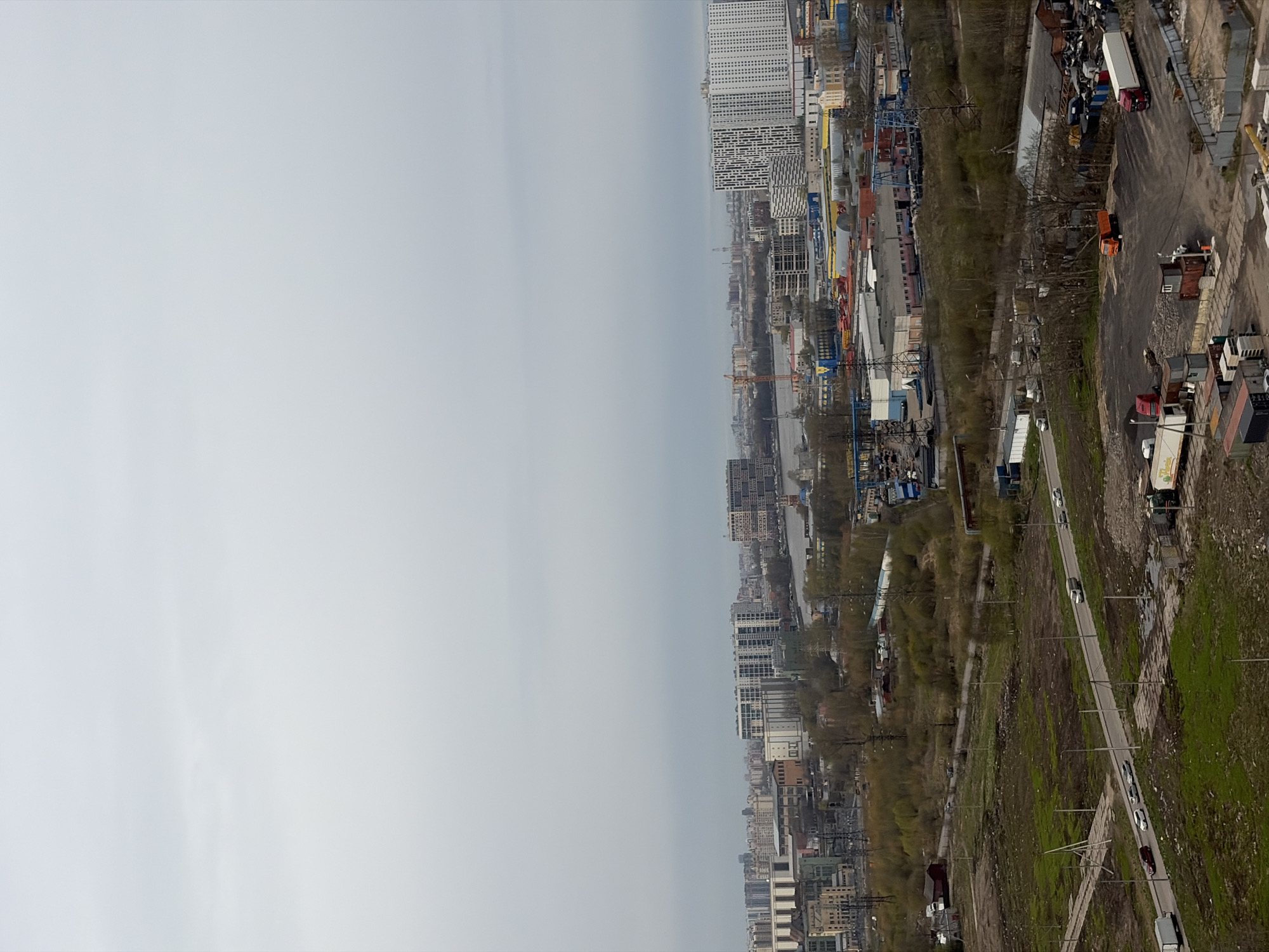 This aerial or high-angle shot captures a sprawling urban and industrial landscape under an overcast sky, characteristic of Saint Petersburg, Russia. The scene is presented from a high vantage point, looking across a vast area that transitions from rugged industrial zones to dense cityscapes.
The foreground is dominated by a raw, unkempt industrial area featuring dirt paths and partially paved roads. Patches of green grass intersperse with sparse, bare trees, and various industrial debris. Numerous vehicles are present, including several semi-trailer trucks (one white with a red cab, another orange), box trucks, and cars parked or driving along the main road. Utility poles with power lines crisscross this area. Indistinct writing or a partial logo is barely visible on the side of a white box truck.
Moving into the mid-ground, the landscape becomes more congested with a mix of low-rise industrial buildings and warehouses. Cranes are visible, indicating potential construction or cargo handling activities. A body of water, likely a river or canal, can be seen winding through the distant urban sprawl.
In the far background, the cityscape rises, featuring numerous multi-story buildings, including modern, tall residential high-rises with distinctive patterns on their facades, signifying dense urban development.
The weather is overcast, with a soft, diffused light bathing the entire scene under a light grey sky, suggesting no direct sunlight. The time of day is daytime. No people are discernible, and no specific event is taking place, offering a static, observational view of this diverse urban and industrial environment.