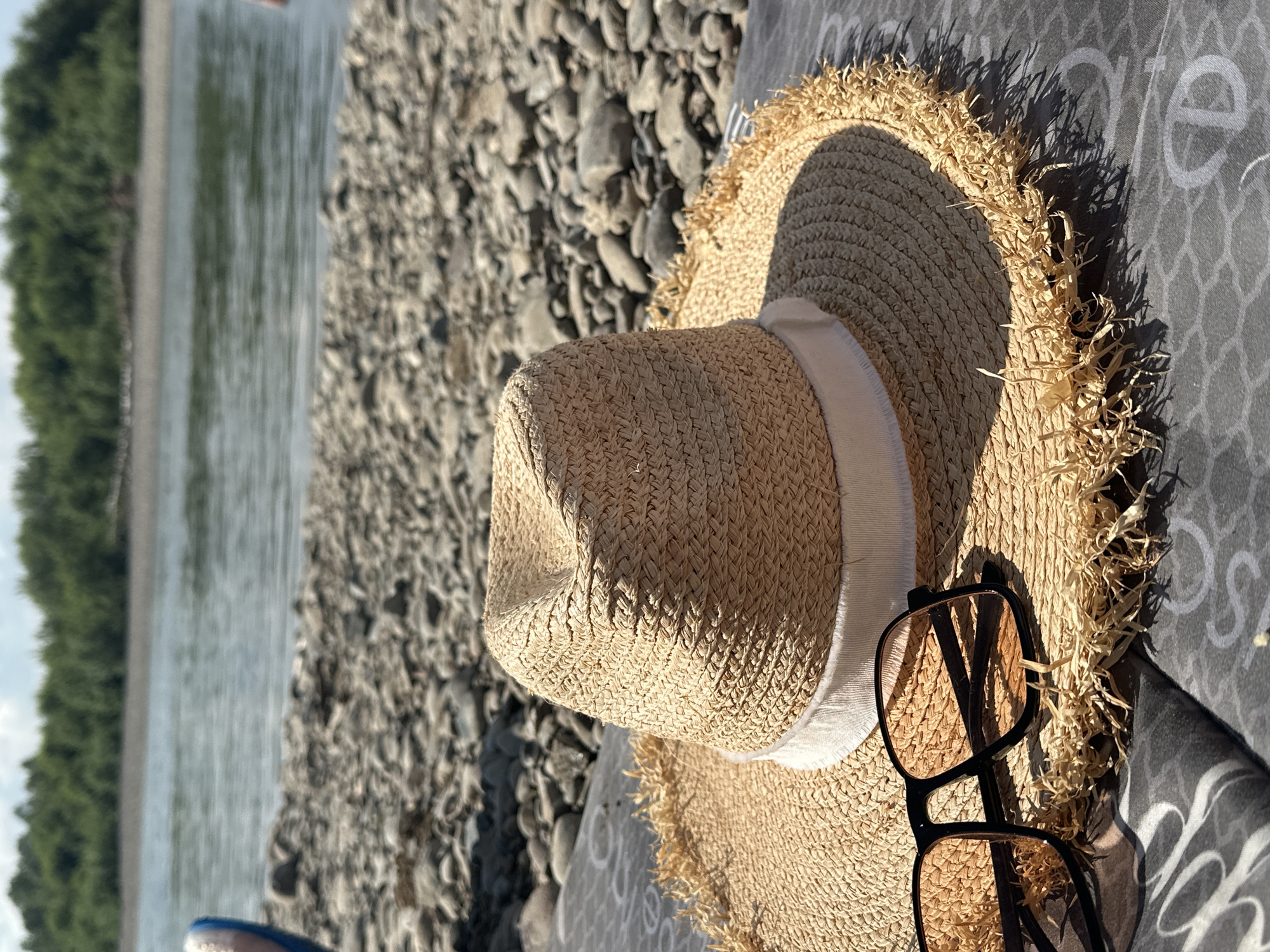 A straw sun hat with a white ribbon band and frayed edges is placed on a patterned blanket on a rocky shore. Beside the hat are a pair of black-rimmed sunglasses with amber lenses. In the background, a calm river flows, with a tree line on the opposite bank. The scene is set in Bely Yar, Russia.