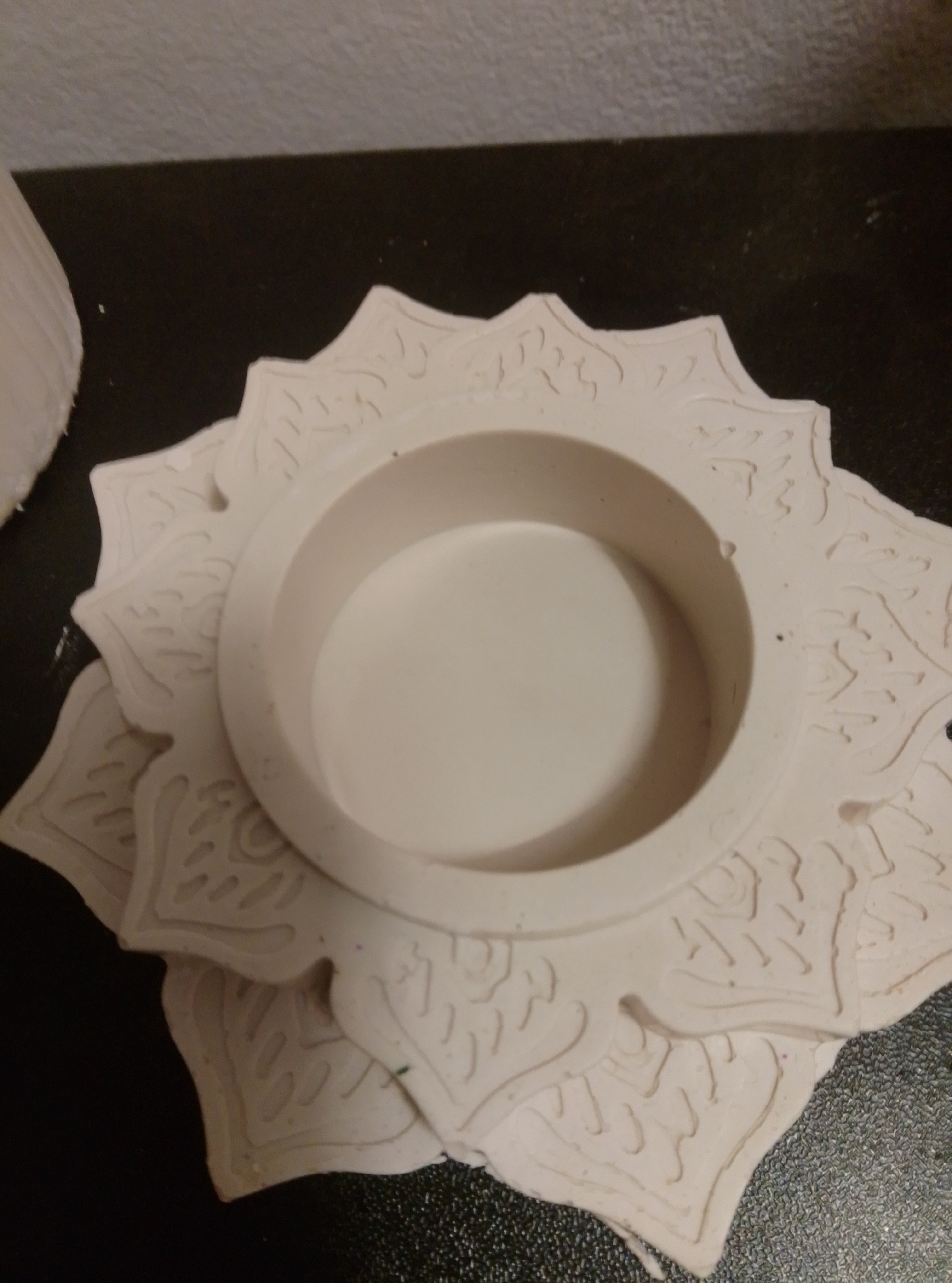 The image shows a white, lotus-shaped ceramic candle holder sitting on a dark, textured surface. The candle holder has intricate embossed patterns on its outer petals. The background is out of focus and appears to be a wall and possibly a piece of white material on the left side of the frame. There are no people visible in the image. The scene is indoors and appears to be a still life. There is no information provided about the location in the city of Wedel, Germany.