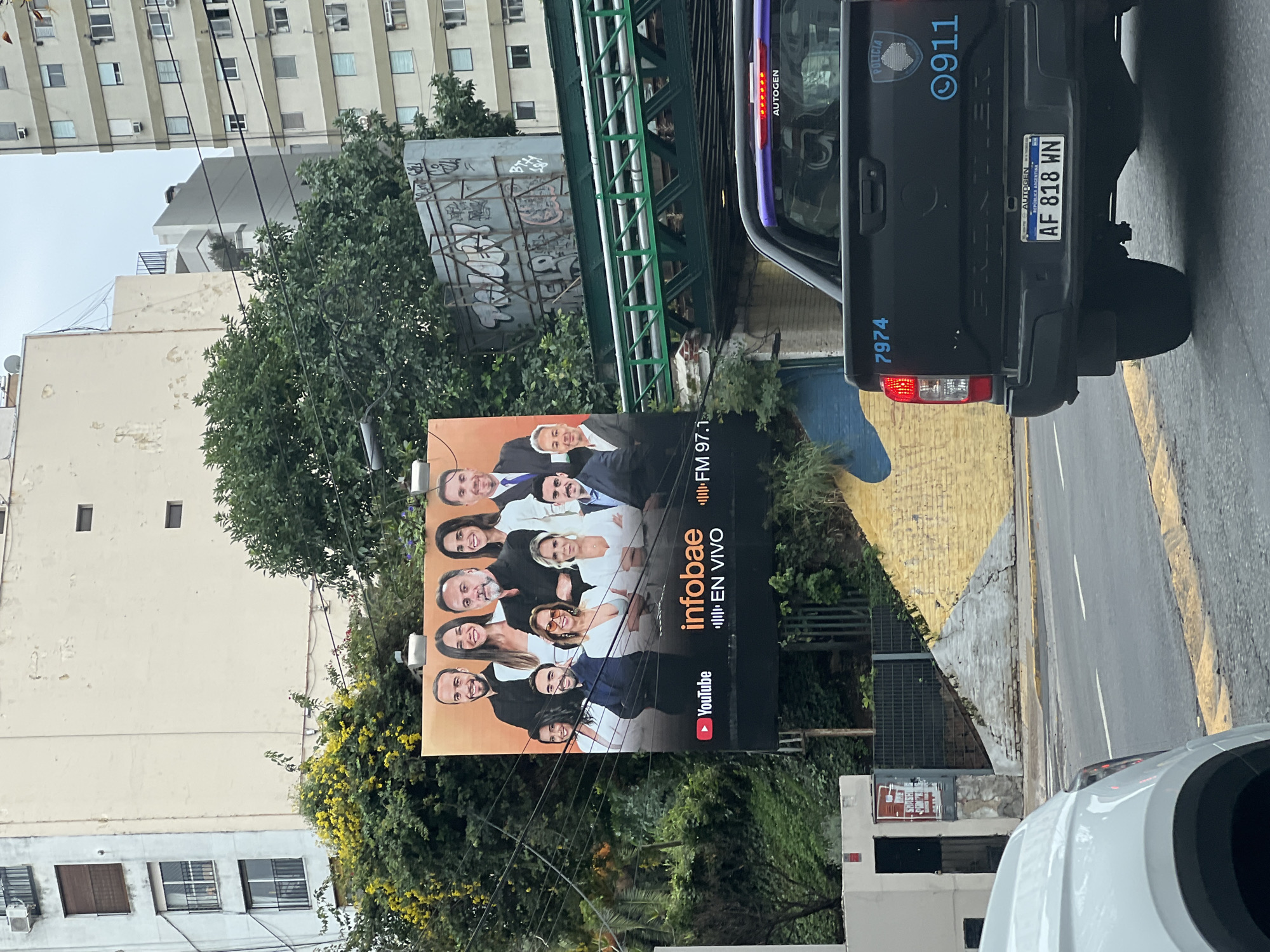 The image shows a street scene in Buenos Aires, Argentina. Prominently displayed is a billboard for "Infobae," a news organization, advertising its FM 97.1 radio channel and YouTube content with an ensemble shot of the Infobae reporters or hosts. To the right of the billboard is a black police truck, displaying the license plate "AF 818 WN," "POLICIA," and the emergency number "911." The truck is partially blocking the view of the billboard. A white car is visible in the lower right corner, further obstructing the view. Behind the billboard and the police truck, there is a bridge and a building covered in graffiti. Buildings of various heights line the street, and trees are scattered throughout the scene. The weather appears overcast.