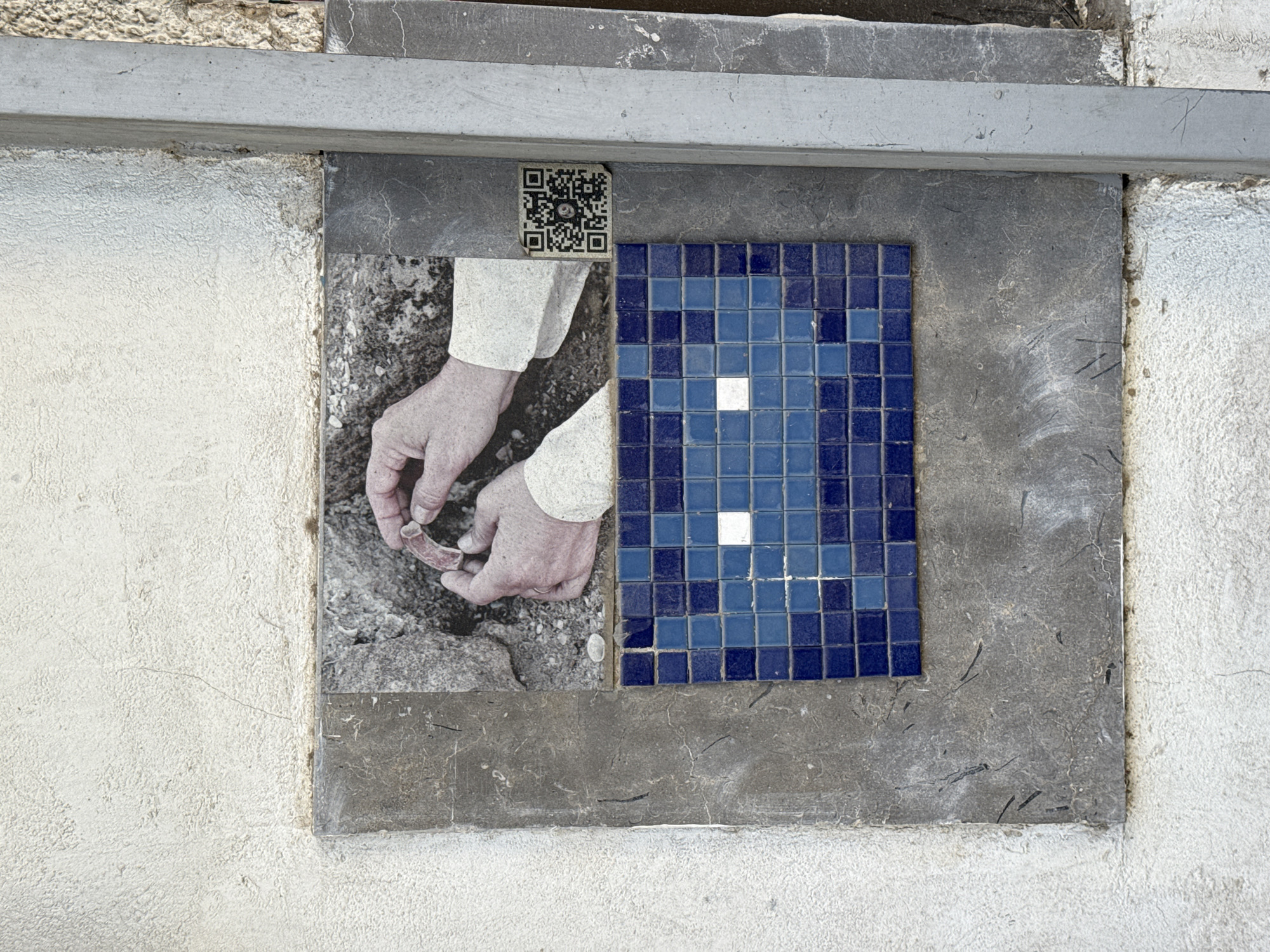 The image shows a piece of street art in Tel Aviv, Israel. It is affixed to a textured white wall, to the right of a vertical metal pipe. The artwork is composed of a rectangular grey stone plaque, on which is mounted a black and white photograph and a mosaic. The photograph, positioned in the upper portion of the plaque, depicts a pair of hands holding a curved, reddish object against a rocky background. A QR code is affixed to the right side of the photograph. Below the photograph is a mosaic of blue square tiles, arranged to form a rectangular shape with two white square tiles in the center, resembling eyes. The overall composition is a stylized pixelated face.