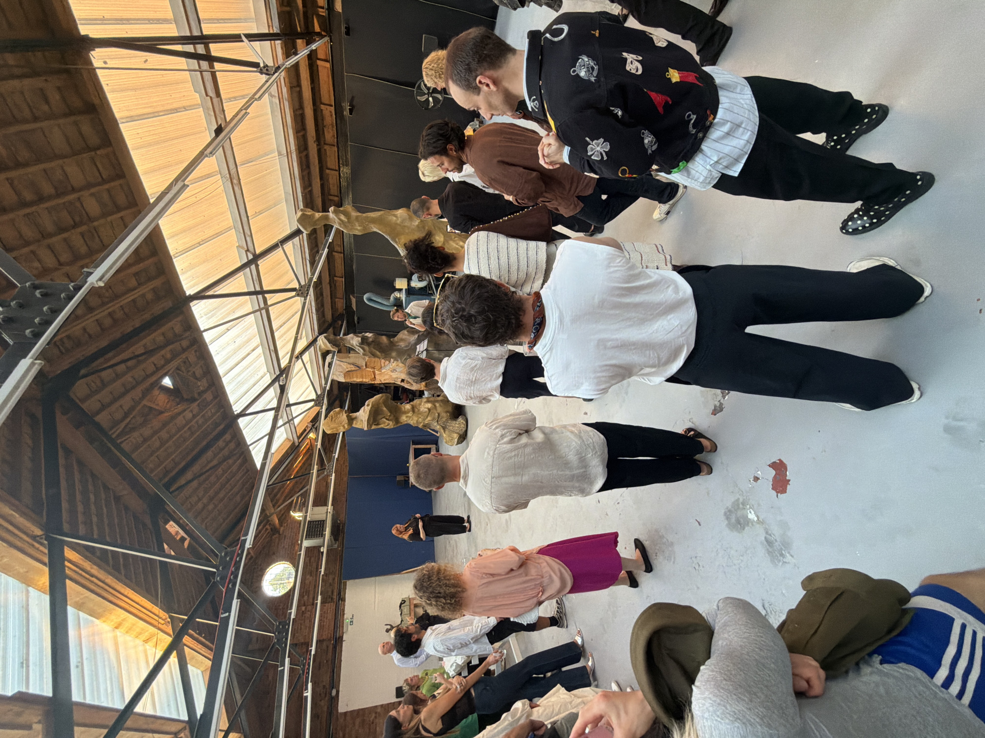 An indoor gathering is taking place in a large, industrial-style space. The ceiling features exposed beams and a translucent, corrugated roof, allowing natural light to filter in. In the background, several abstract, golden sculptures are positioned. A group of approximately fifteen people, of varying ages and ethnicities, are present, some standing and observing the sculptures, while others appear to be engaged in conversation. One woman in the center, wearing black clothing, is speaking to the group. The floor is a light grey concrete, showing some wear and tear. The overall atmosphere suggests an art exhibition or a similar cultural event. The location is not identifiable from the image alone.