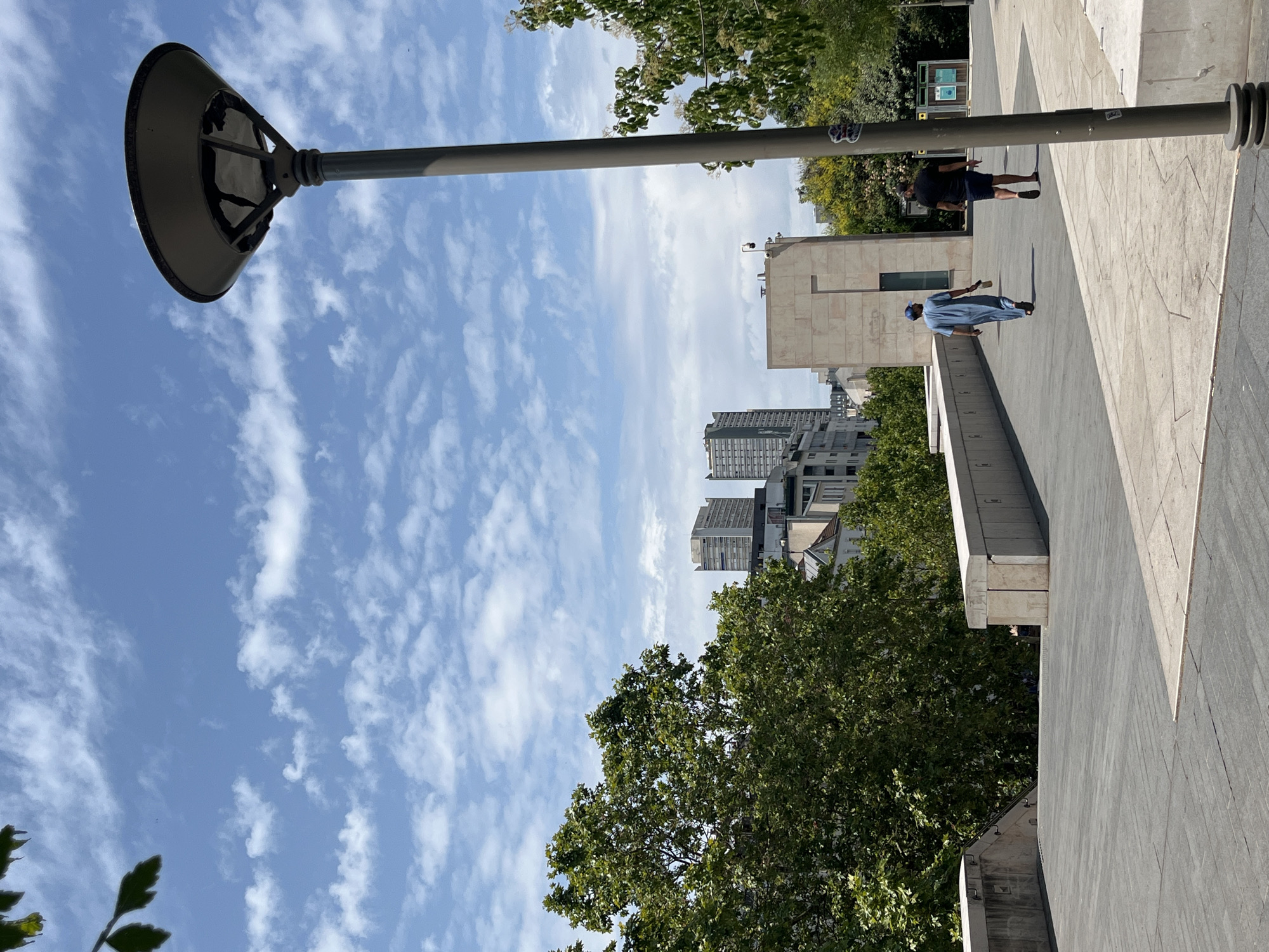 The image shows a partially rotated view of an outdoor urban setting on a sunny day with a bright blue sky scattered with white clouds. A large, dark, circular lamp post head is prominent in the upper left, extending horizontally across the frame. The lamppost itself is a thick, gray pole. In the middle ground, several people are visible on a wide, paved walkway. One person, wearing a light blue garment and a cap, is walking away from the camera, holding a dark object. Another person, wearing a dark shirt and shorts, stands with their back to the camera, facing the direction of the walking person. Behind them, a modern building with large, light-colored rectangular panels forms a prominent architectural feature. Further in the background, a cluster of taller buildings, typical of a city skyline, can be seen. Lush green trees frame the scene, particularly in the lower right and upper right portions of the image, suggesting a park or a tree-lined street. The sun casts distinct shadows, indicating it is likely daytime, possibly late morning or early afternoon. The overall impression is one of a pleasant, ordinary day in a city, with people engaged in casual movement.