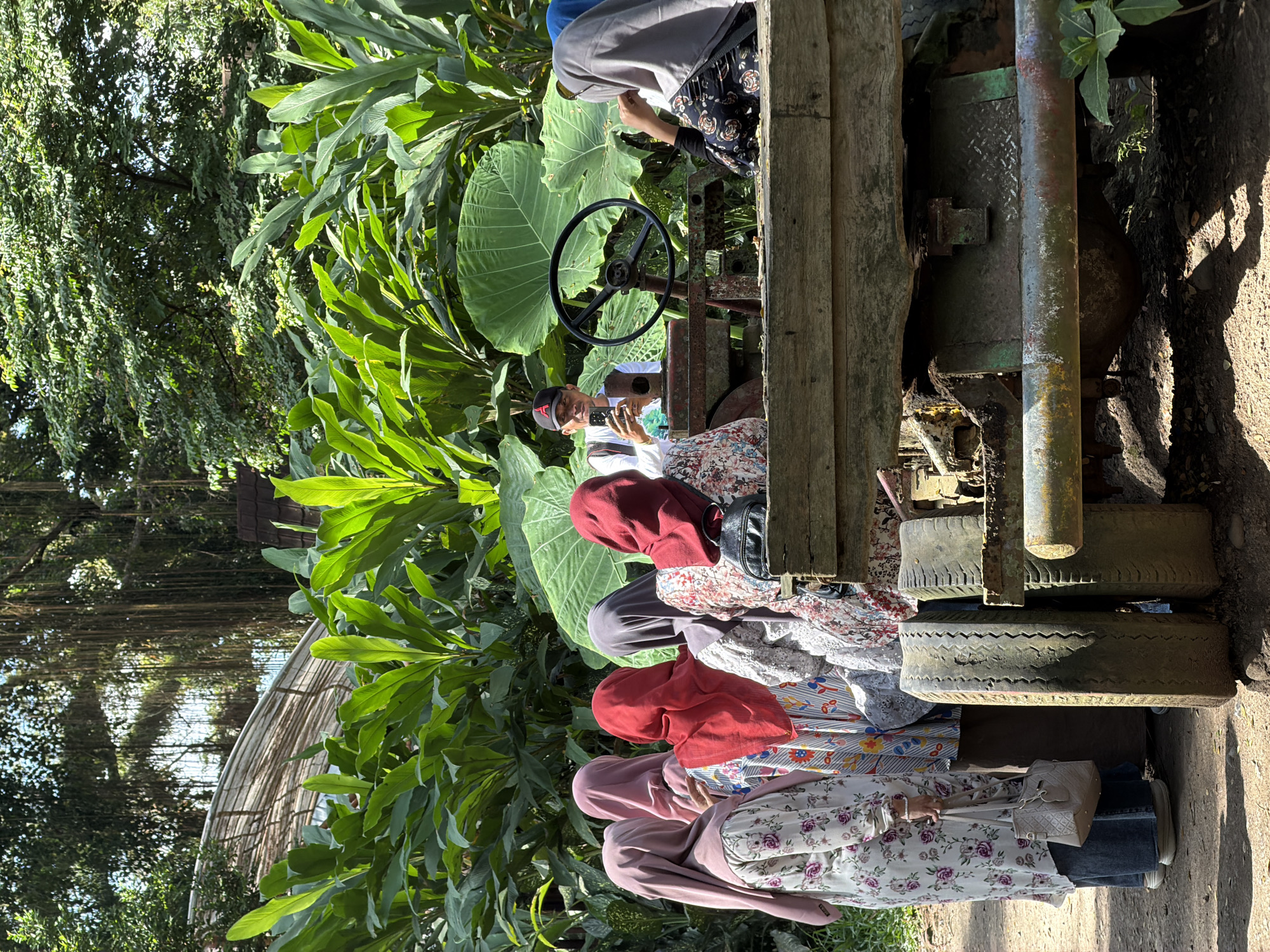 In Sraten, Indonesia, a group of individuals, predominantly women wearing hijabs, are gathered around a weathered, wooden cart with visible metal components and tires. One person, a man wearing a baseball cap, is taking a photograph with his phone, while another woman in a grey hijab appears to be looking in his direction. The cart is situated outdoors, amidst lush green foliage, with large leaves prominently featured. The background suggests a natural, possibly tropical environment, with trees and hanging vines visible. The lighting indicates it is daytime, with sunlight casting shadows.