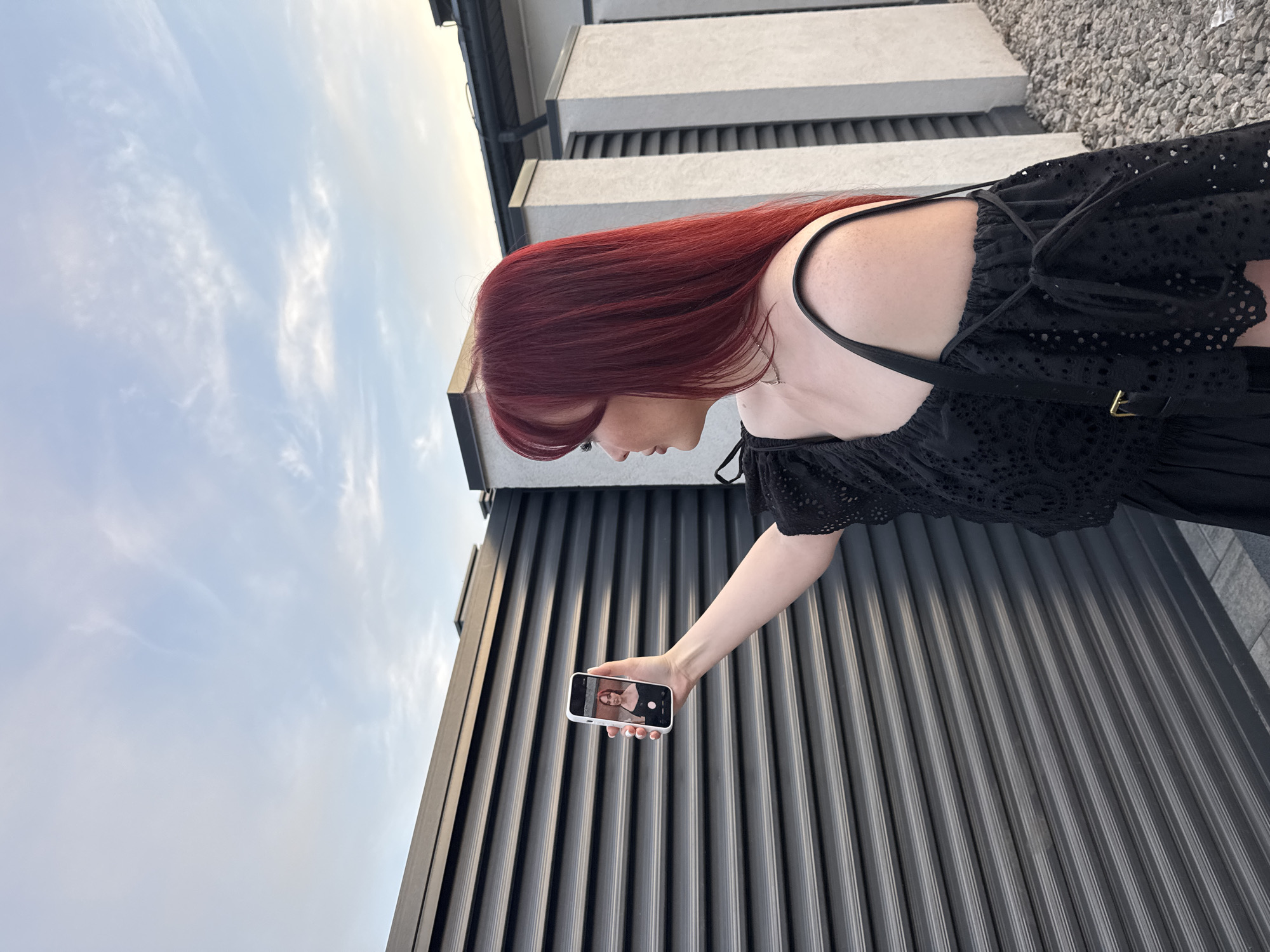 A woman with long, red hair stands outdoors, angled away from the camera, taking a photo with her smartphone. She wears a black dress with shoulder straps. The phone screen displays a picture of a person. Behind her is a wall with vertical lines, and above that, a building with grey horizontal lines and a small area covered with small stones. The sky is visible in the upper left corner with sparse clouds. The location is Unknown, Unknown.