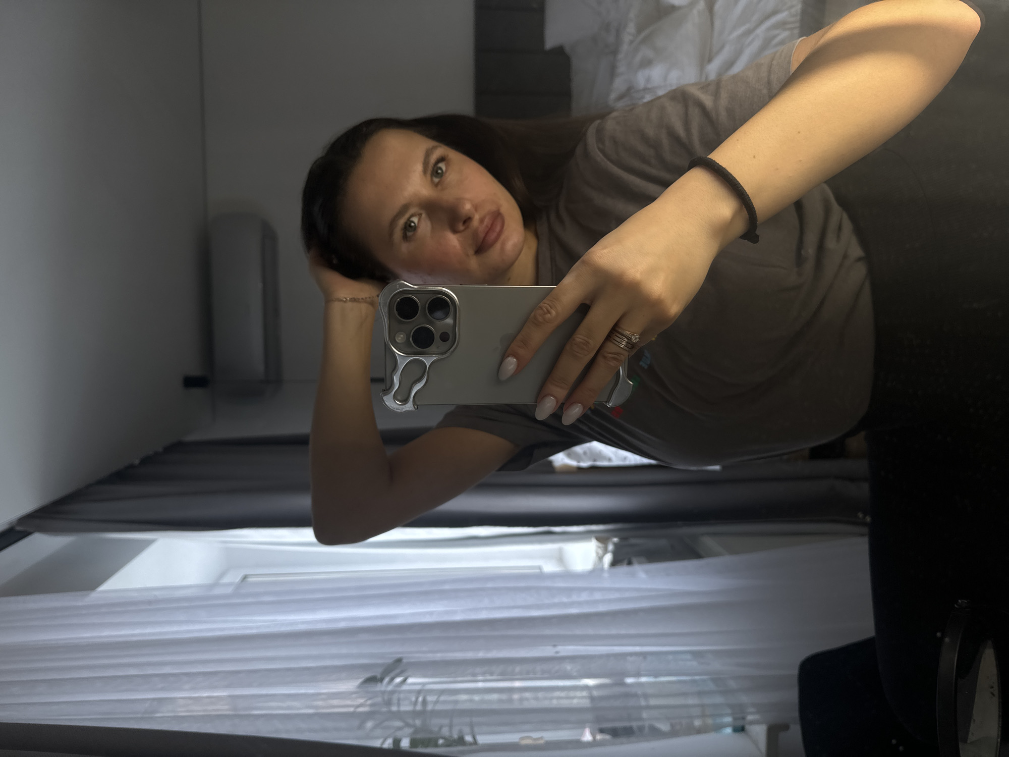 The image shows a woman taking a mirror selfie in an indoor setting. The photograph is oriented vertically, with the woman's body appearing horizontal across the frame. She has long, dark hair and is wearing a plain grey t-shirt. Her right arm is bent, with her hand resting behind her head, and a thin bracelet is visible on her wrist. Her left hand holds a silver smartphone with a clear or silver-toned protective case, which she is using to capture the reflection. Rings, including what appears to be a wedding band, are visible on her left hand, and her fingernails are painted with light polish.
She is looking directly into the reflected camera lens with a neutral to slightly soft expression. The background, visible in the mirror's reflection, indicates an indoor space, possibly a bedroom or hotel room. Part of a bed with white bedding is visible in the upper right portion of the reflection. On the left, a light grey wall or cabinet is reflected. In the lower area of the reflection, horizontal window blinds or shades can be seen, suggesting a window. A dark object, potentially a piece of furniture, is partially visible on the far right of the reflection. The lighting is consistent with indoor illumination. No text is visible in the image.