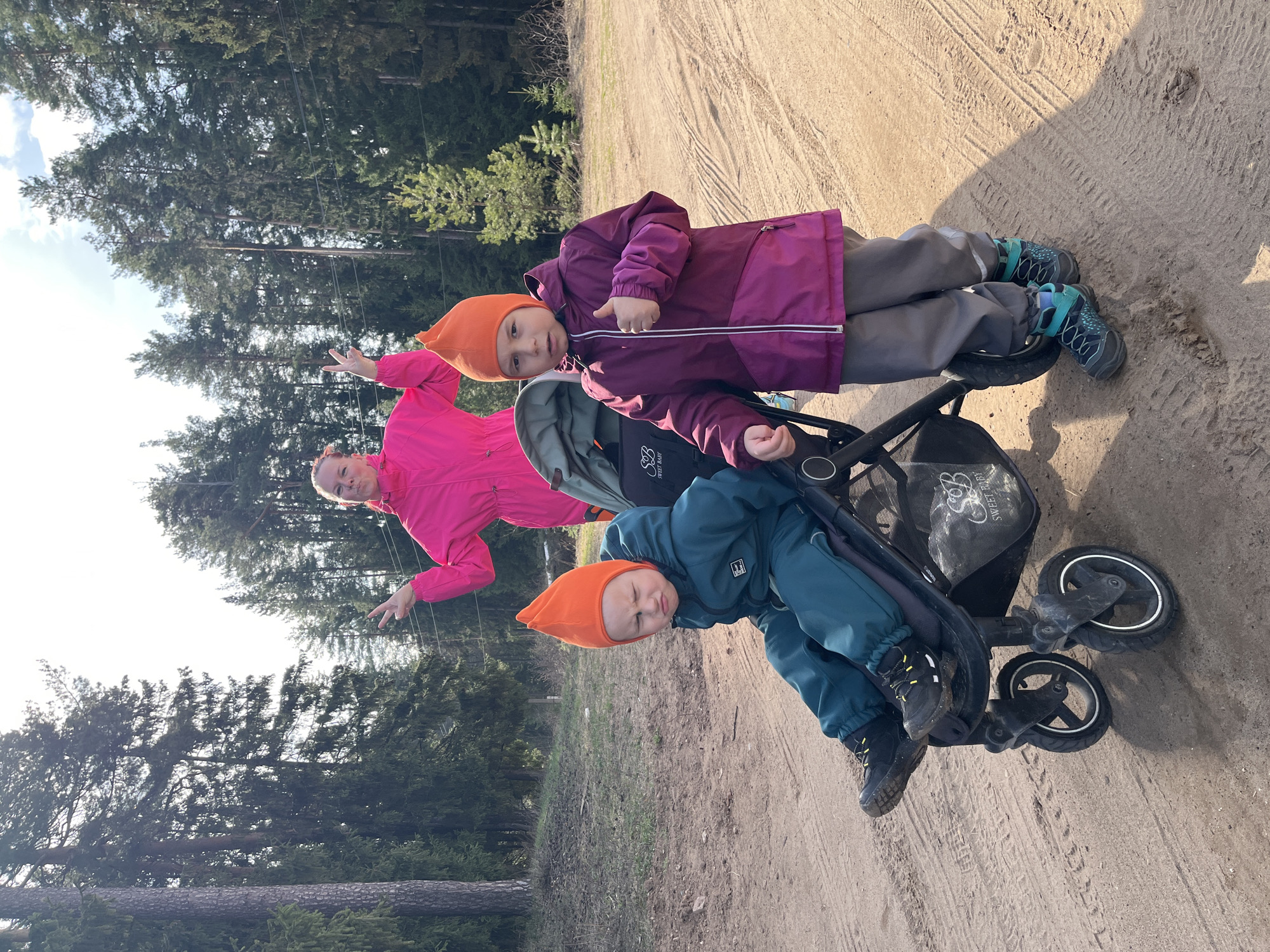 In an outdoor setting near a forest in Chaikovsky, Russia, two children wearing orange cone-shaped hats are positioned in a stroller on a sandy path. The child sitting in the lower part of the stroller is wearing a teal blue snowsuit and dark shoes. The child positioned above is wearing a purple snowsuit, gray pants, and teal-accented hiking boots. A woman wearing a bright pink jacket stands to the side, partially obscured by the trees. She has both of her arms extended with an open palm gesture and is smiling at the camera. The stroller has a black mesh bag with the text "Sweet Baby" visible. The ground is covered in sand, and tire tracks are apparent. Behind the group, the forest has tall evergreen trees. The image is taken during the daytime with natural lighting.