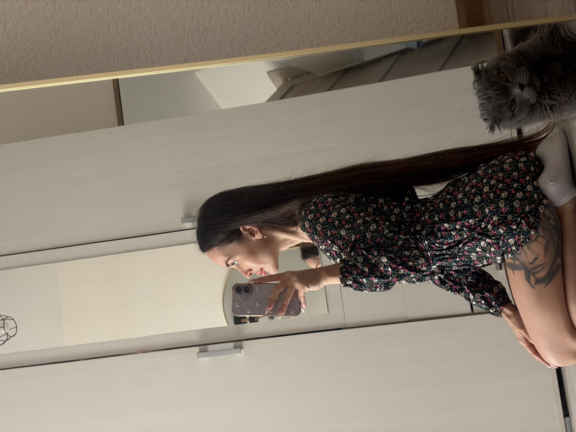 The image displays a mirror selfie taken by a young woman indoors, presumably in Saint Petersburg, Russia. The original image is rotated 90 degrees clockwise.
The main subject is a woman with long, straight dark hair, dressed in a dark floral mini-dress featuring pink, white, and green patterns. She is captured in profile, intently looking at her light-colored smartphone, which has a decorated case, as she holds it up to take the photo. Small earrings are visible on her ears, and she has visible freckles or minor blemishes on her face. A large tattoo of a woman's face is prominent on her upper left thigh or hip. Her feet are seen wearing white socks or slippers.
In the upper right portion of the mirror's reflection, a fluffy grey cat with amber eyes is visible, looking directly towards the woman or the camera. The cat appears to be situated on a surface above the woman.
The setting appears to be a bedroom or dressing area. The mirror has a thin, golden-colored frame at its top edge. Below the mirror, a light-colored (possibly white or off-white) wardrobe or dresser with two sections and silver horizontal handles is visible. Above the mirror, a textured light brown or beige wallpaper covers the wall. The lighting is soft and consistent with indoor artificial light. No visible text is legible in the image.