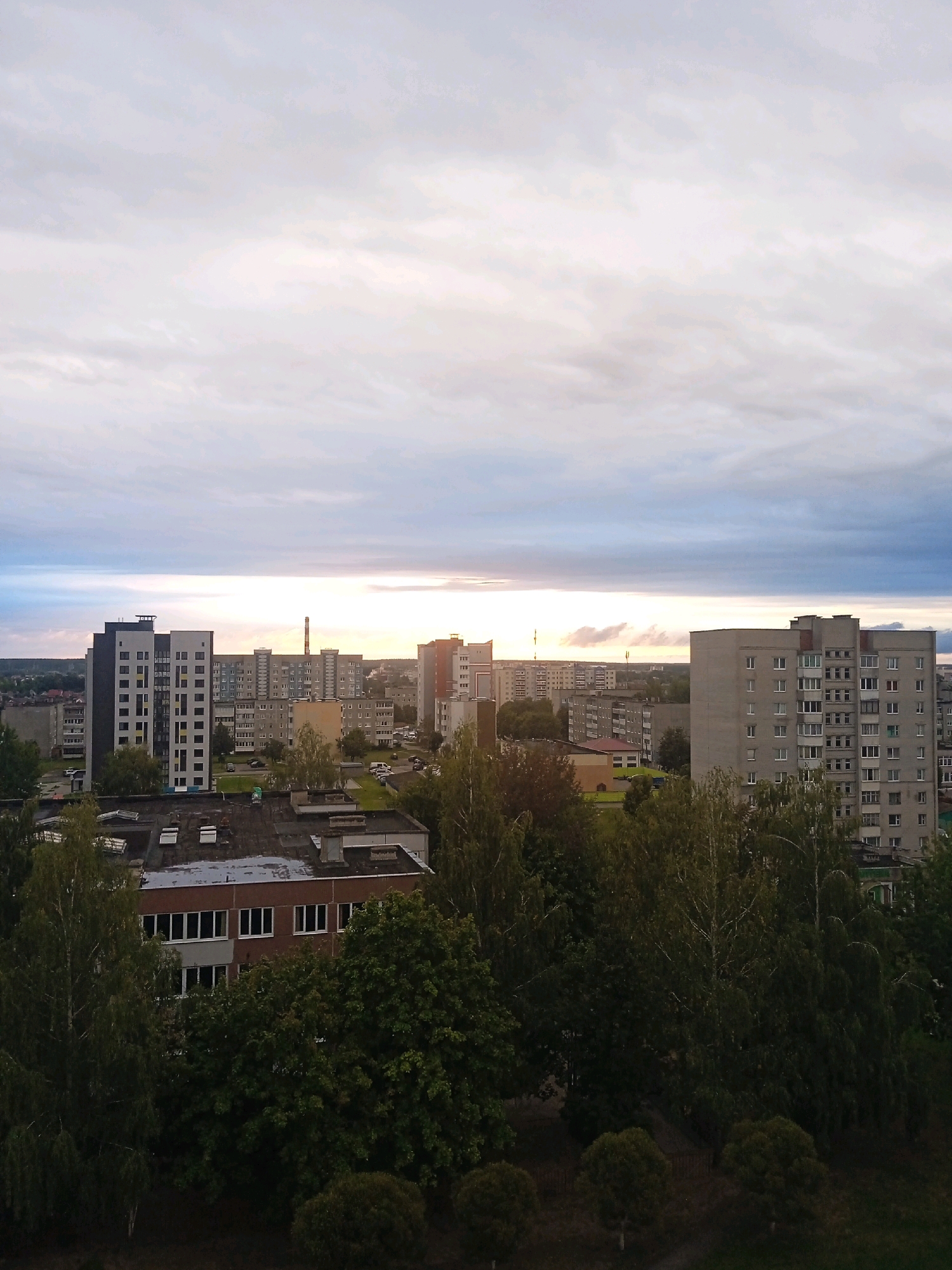 This is an aerial photograph of a city skyline with a cloudy sky. In the foreground, a dark, tree-lined embankment is visible, with the dark roofs of lower buildings peeking through the foliage. Behind this, several multi-story apartment buildings and other urban structures are visible, varying in height and architectural style. A prominent tall, slender building with a red-capped chimney or antenna is visible in the mid-ground. The sky above is filled with a mix of white and grey clouds, with a bright band of light on the horizon suggesting a sunset or sunrise. The scene appears to be residential or mixed-use urban development.