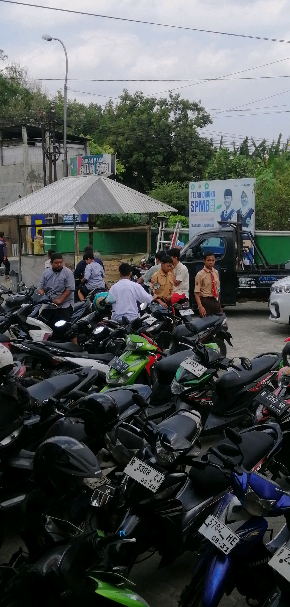 The image depicts an outdoor scene in Windunegara, Indonesia, likely a street or parking area. In the foreground, a dense collection of parked motorcycles fills the frame, with several individuals seated on or standing near them. The people appear to be a mix of adults and young people, some in casual attire and others in school uniforms, suggesting a possible school event or gathering.
Behind the motorcycles, a small, open-air food stall with a corrugated metal roof is visible. A sign above it reads "RUMAH MAKAN AULIA SARI." Further back, a dark pickup truck is parked, and to its right, a large banner announces "TELAH DIBUKA SPMB 2020" (SPMB 2020 is open), featuring images of two individuals in school uniforms. The background consists of trees and a cloudy sky. The overall impression is a bustling, everyday scene in an Indonesian urban or semi-urban setting.