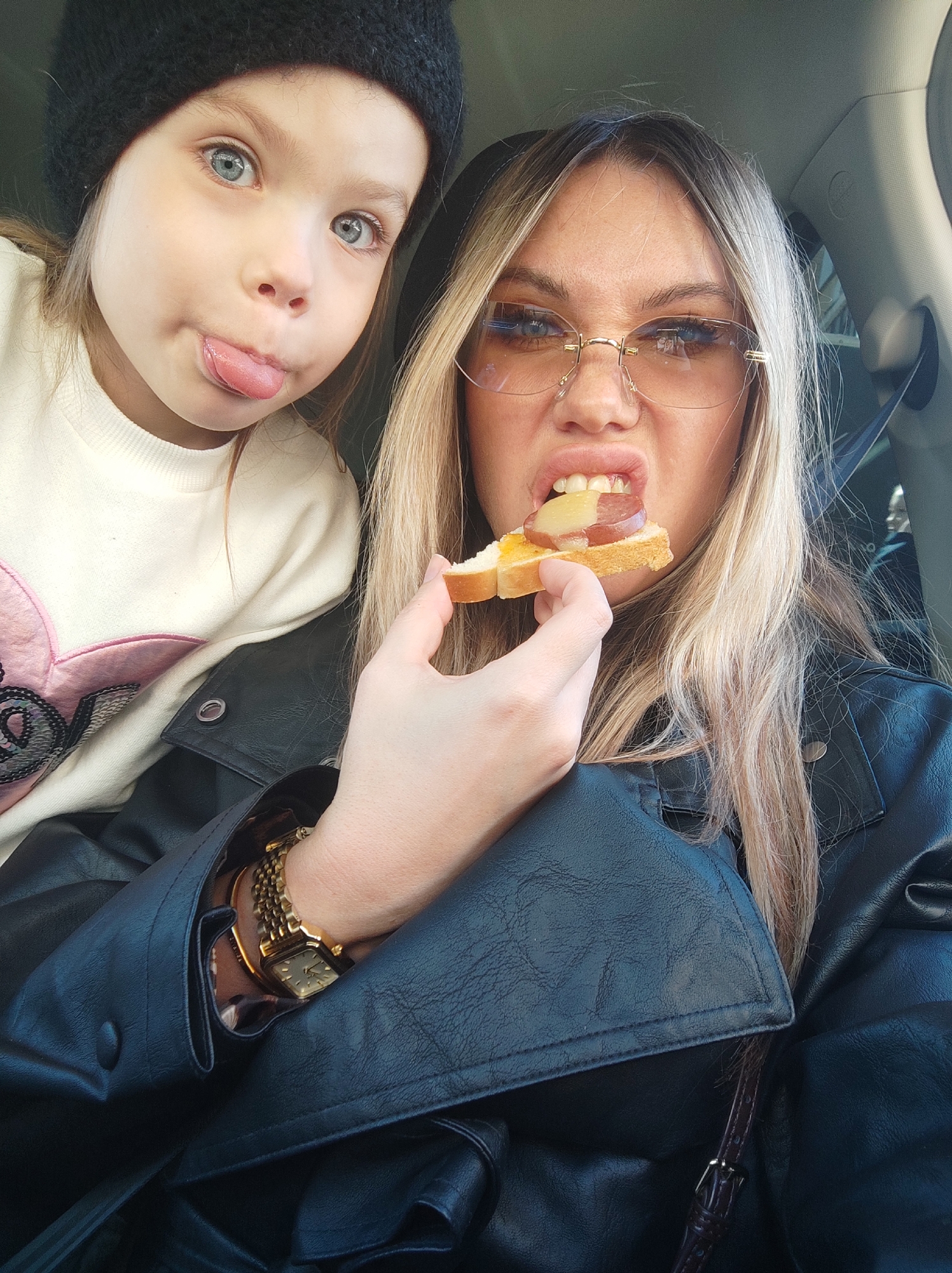 The image captures a woman and a young girl, likely in the interior of a car, potentially in Izhevsk, Russia (though this cannot be definitively confirmed without additional context). The girl, wearing a dark knit cap and a light-colored sweater with a heart design, is sticking her tongue out. The woman, beside her, is wearing glasses and a black leather jacket, and she's holding a piece of bread topped with what appears to be sausage and a slice of apple or pear, partially in her mouth. Her hair is blonde with darker roots. She also appears to be wearing a gold watch on her left wrist. The vehicle's interior features are visible in the background, with some of the car's structural elements distinguishable.