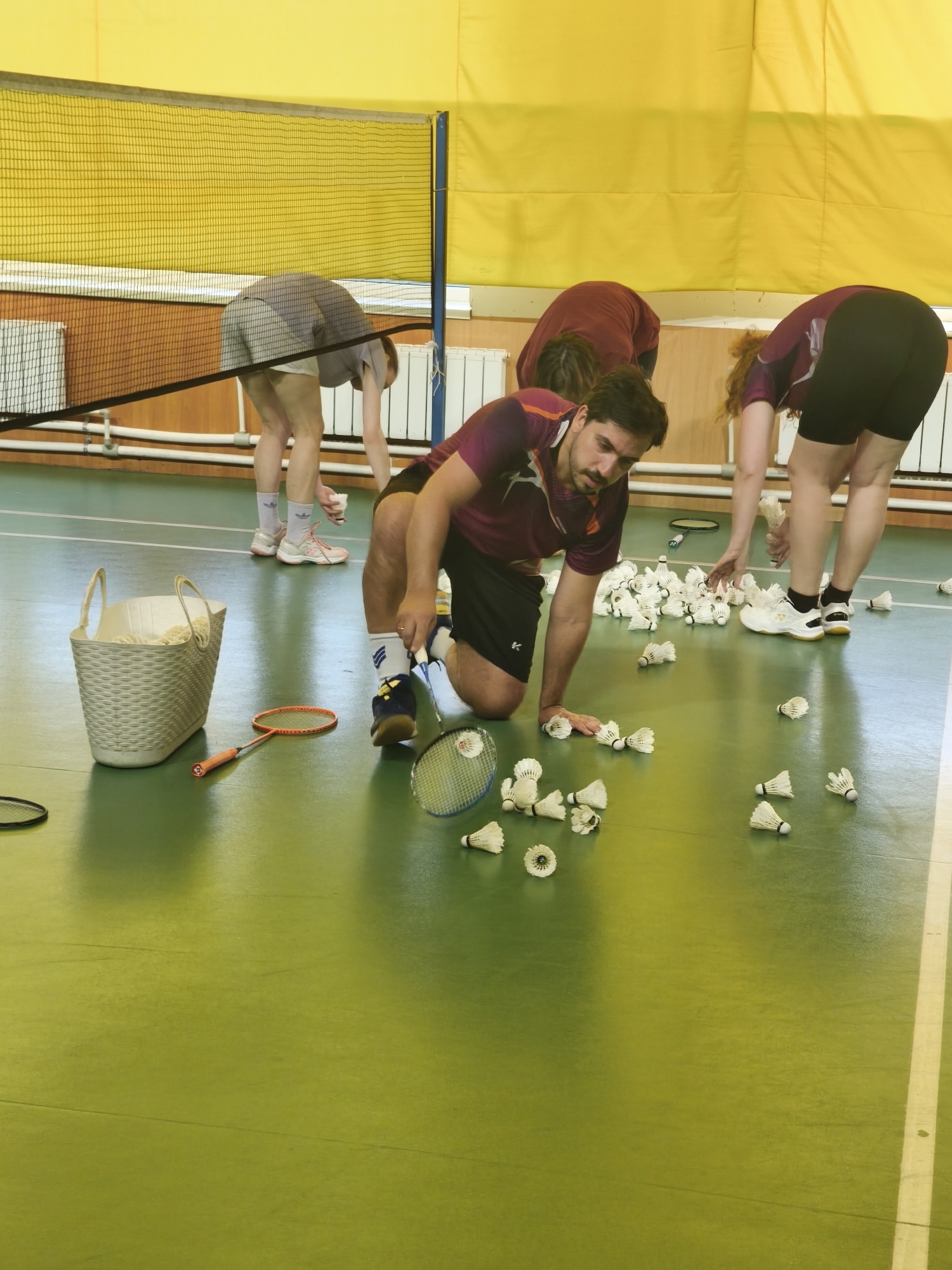 An indoor sports court features four individuals engaged in collecting numerous white shuttlecocks scattered across a vibrant green floor. The court is marked with faint white lines. A black badminton net with a yellow top band extends across the mid-ground.
In the immediate foreground, a male with fair skin, dark hair, a beard, and mustache is kneeling on his right knee. He wears a maroon and orange athletic shirt, black shorts with a white logo on the left thigh, blue and white athletic shoes with blue laces, and white socks with blue stripes. His right hand is reaching down to collect a shuttlecock. A blue and white badminton racket lies partially visible beside him.
To his right, a female with reddish-brown curly hair, wearing a similar maroon athletic shirt, black shorts, white athletic shoes with yellow accents, and black socks, is bent forward, her hands gathering a pile of shuttlecocks.
In the mid-ground to the left of the net, another individual, partially obscured, is bent over, wearing a light grey t-shirt and shorts, with white and pink athletic shoes and light purple socks. They are also picking up shuttlecocks. Behind the net, a fourth person, visible from the back, is bent forward, wearing a dark red long-sleeved shirt.
On the left side of the court, near the foreground, a light beige plastic woven basket with handles rests on the floor, containing white string-like material. An orange badminton racket with a black grip lies next to it.
The background wall features a lower section of light brown wood paneling. Above this, white horizontal pipes and radiators are mounted. The upper section of the wall is covered with a yellow padded fabric or curtain. A horizontal light-colored opening is visible on the upper left wall.
The overall scene depicts a clean, well-lit indoor sports facility where individuals are systematically gathering shuttlecocks. The scene is located in the city of Saint Petersburg, Russia.