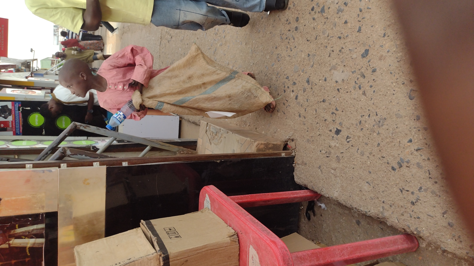 A pedestrian carrying a sack walks on a concrete surface in Jos, Nigeria. They are wearing a peach-colored long-sleeved shirt and appear to be carrying goods in a large sack. To their left is a temporary stall with boxes and a red plastic chair. Other people are visible in the background.