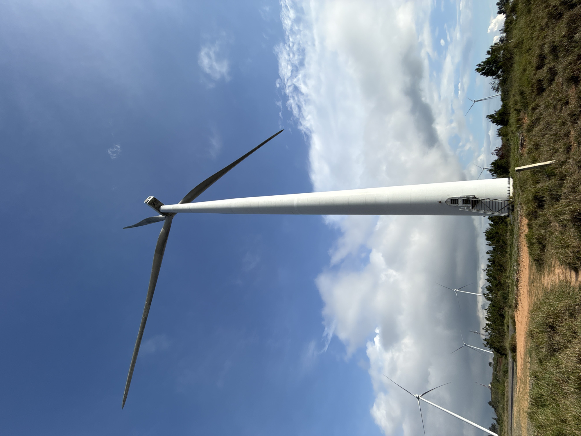 The image shows a wind turbine in Phát Chi, Vietnam. The turbine is white and has three blades. It is located in a field with other turbines visible in the background. The sky is blue with scattered clouds. The ground is covered in grass and vegetation.