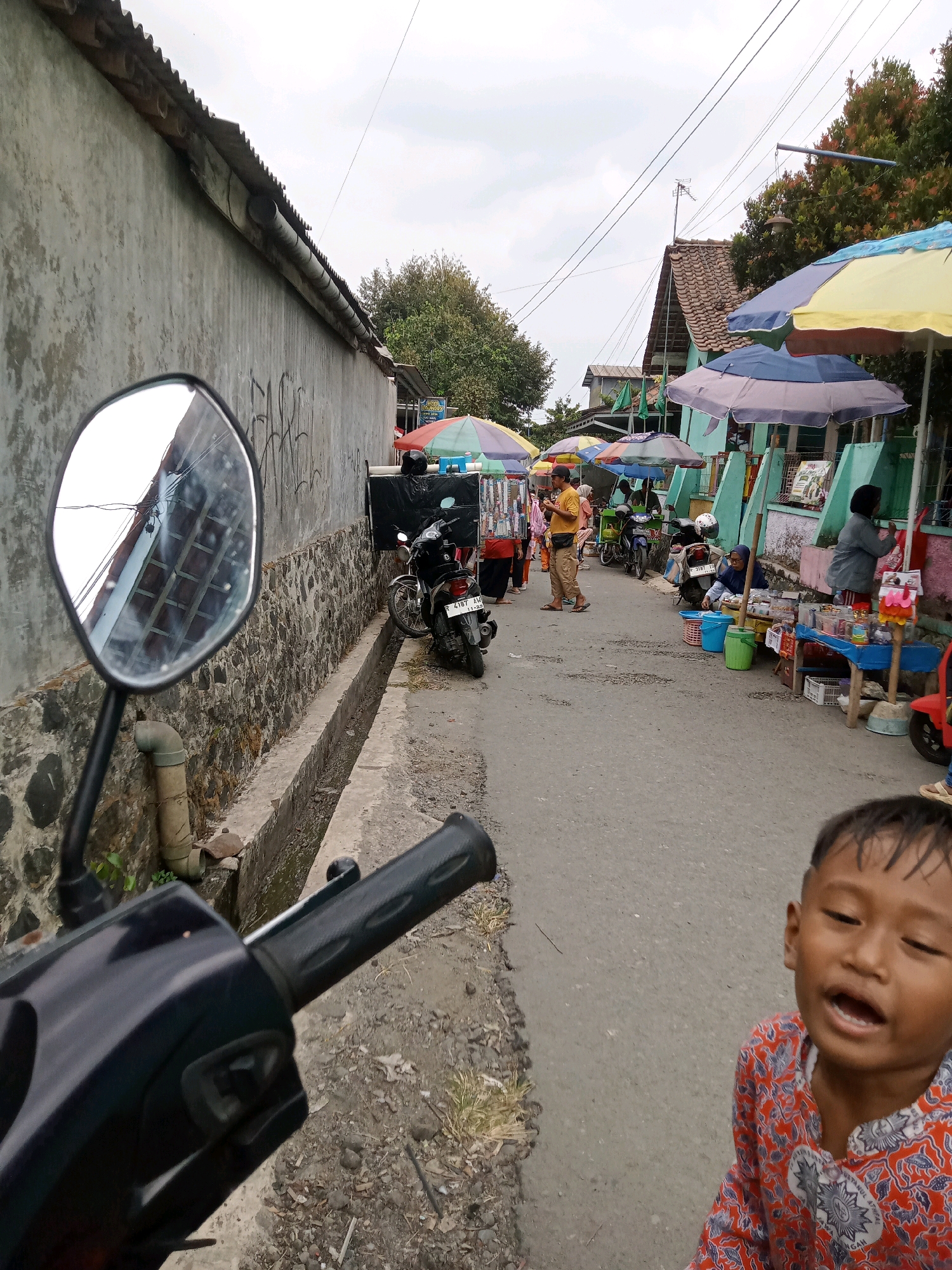 The image depicts a street scene in Gumiwang, Indonesia. The foreground features the handlebar and side mirror of a dark-colored scooter. The mirror reflects a tiled roof and a stone wall. To the right of the scooter, a young boy with short hair, wearing a red and white patterned shirt, looks towards the camera with his mouth open.
The street is lined with market stalls covered by colorful umbrellas. Several people are visible around the stalls, some appearing to be vendors and others customers. One stall features merchandise displayed on tables, with buckets and baskets underneath. A black motorcycle is parked near one of the stalls. The street itself is paved and appears to be a pathway or narrow road. A drainage channel runs along the left side of the street, separating it from the stone wall. Trees and buildings are visible in the background, suggesting a residential or commercial area. The sky is overcast.