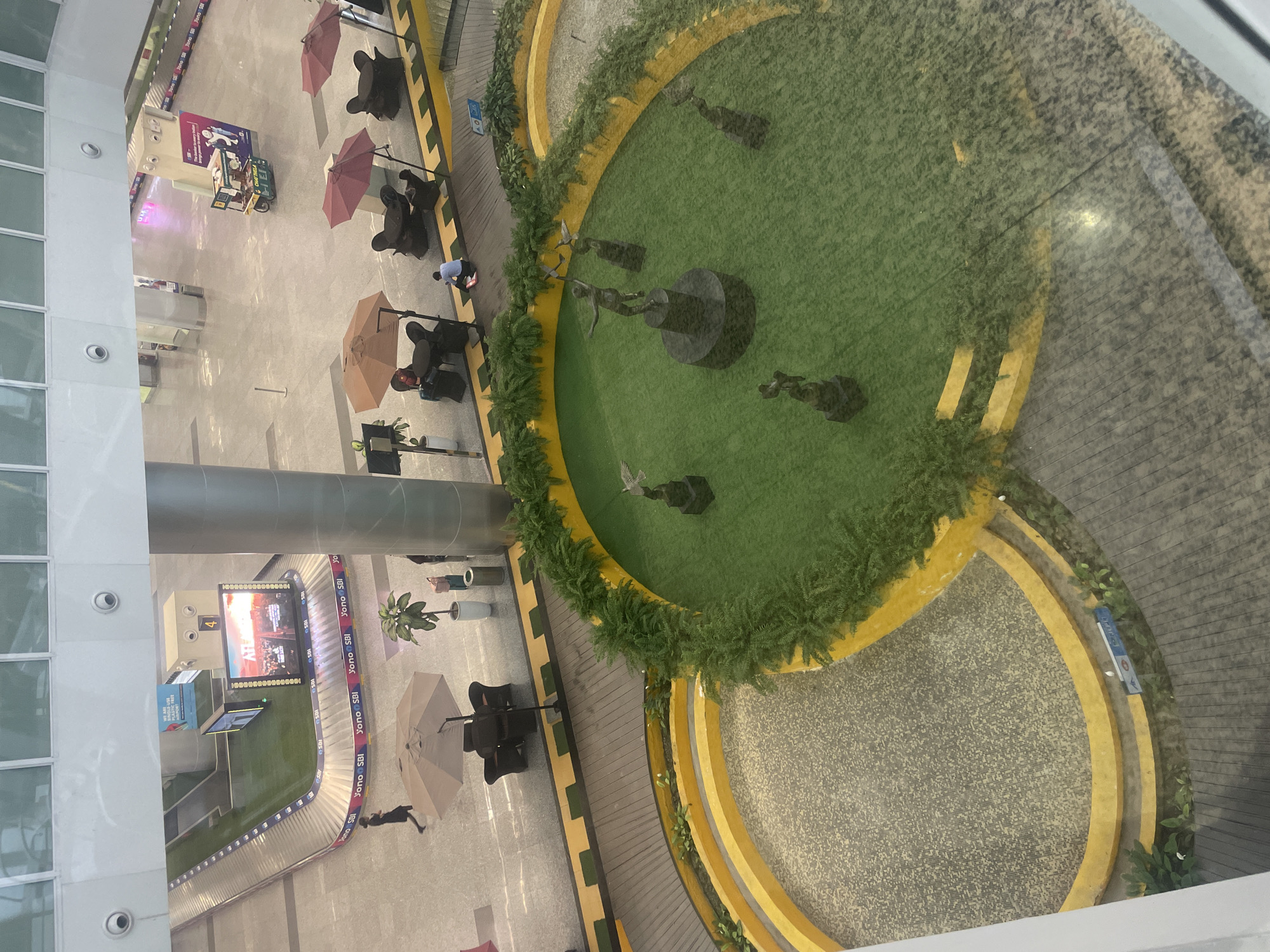 The image depicts the interior of an airport terminal, likely in Raipur Khurd, India, as suggested by the presence of a baggage claim area with signage referencing "Yono SBI." The scene is viewed from an elevated perspective.
In the foreground, a circular, green area is visible, resembling artificial grass, which serves as a decorative centerpiece. Within this green space, several bronze sculptures are strategically placed. One prominent sculpture depicts a figure with outstretched arms, appearing to be in motion or celebratory. Other sculptures appear to be smaller figures, possibly birds or stylized human forms, also on pedestals. The green area is bordered by a yellow, curved structure and lush green plants.
To the left of the central green area is a curved, textured surface, likely a decorative flooring material, also framed by yellow borders. Beyond this, a wooden walkway is visible.
In the mid-ground, several seating areas with umbrellas and dark-colored chairs are arranged. A few individuals are present, with one person seated and another walking. A potted plant and a trash receptacle are also noted in this area.
Further back, the baggage claim area is clearly visible, with a conveyor belt system in place. A large digital display screen shows an advertisement for "ATLANTIS" with a cityscape image. Another kiosk-like structure with signage including the number "4" is also present.
The overall environment is a modern airport terminal with polished floors, metallic columns, and overhead lighting. The perspective suggests the photograph was taken from a higher level within the terminal. The presence of a baggage claim area, advertising screens, and seating indicates this is a public transit hub.
