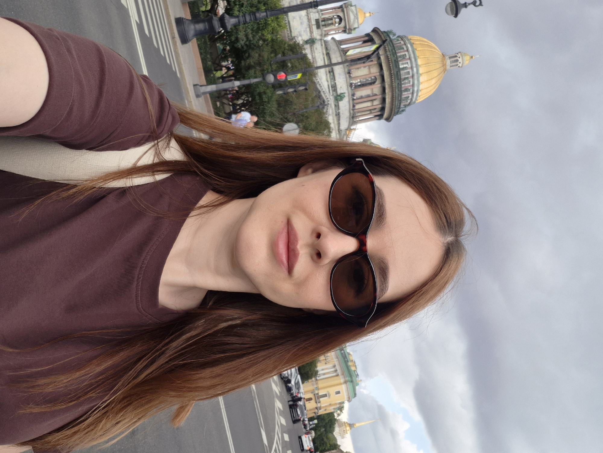A woman with long auburn hair is taking a selfie in front of a large domed building with a golden dome. She is wearing sunglasses and a brown t-shirt. Behind her, there is a street with cars and other buildings. The sky is cloudy.