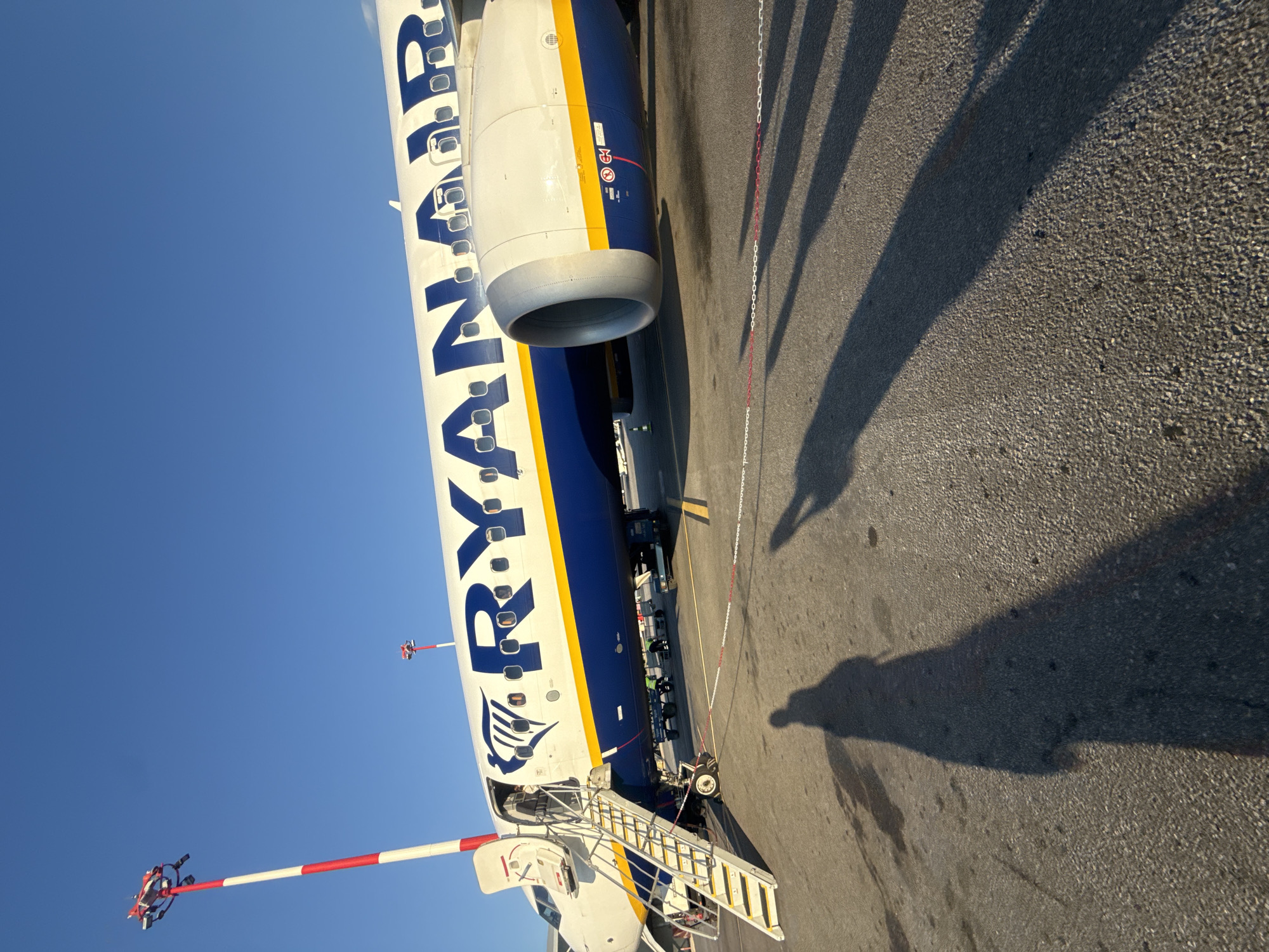 An outdoor, medium-shot, eye-level, wide-angle photograph captures a Ryanair airplane parked on an asphalt tarmac under a clear blue sky. The airplane is primarily white with a large blue Ryanair logo emblazoned across its fuselage, accented by a yellow stripe beneath the windows. The aircraft's front boarding stairs are extended, with several passengers visible ascending. The plane's engine is prominent in the foreground, and a red and white striped pole, likely a lighting or signal mast, stands to the left of the aircraft. Two red and white ropes delineate a boundary on the tarmac. The shadows of people are cast on the ground, indicating a sunny day. The location is the Thessaloniki Airport (SKTH), also known as Macedonia International Airport, situated in the municipality of Thermi, Greece.