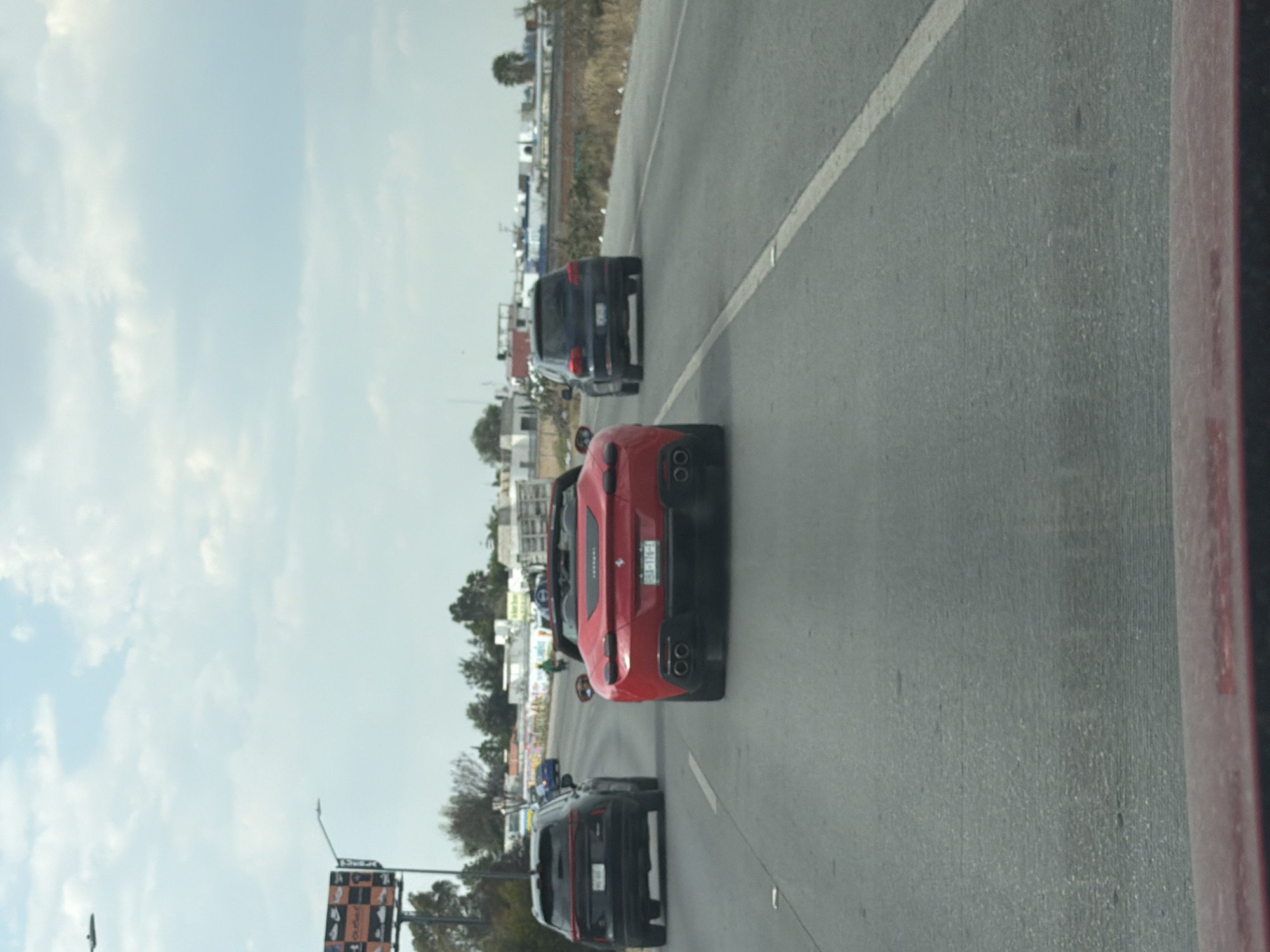 The image shows a line of cars on a highway in San Bernardino Tlaxcalancingo, Mexico. The red Ferrari is the most prominent object in the image, followed by two black SUVs. The highway is made of gray asphalt and has white lane markings. In the background, there are buildings, trees, and a cloudy sky.