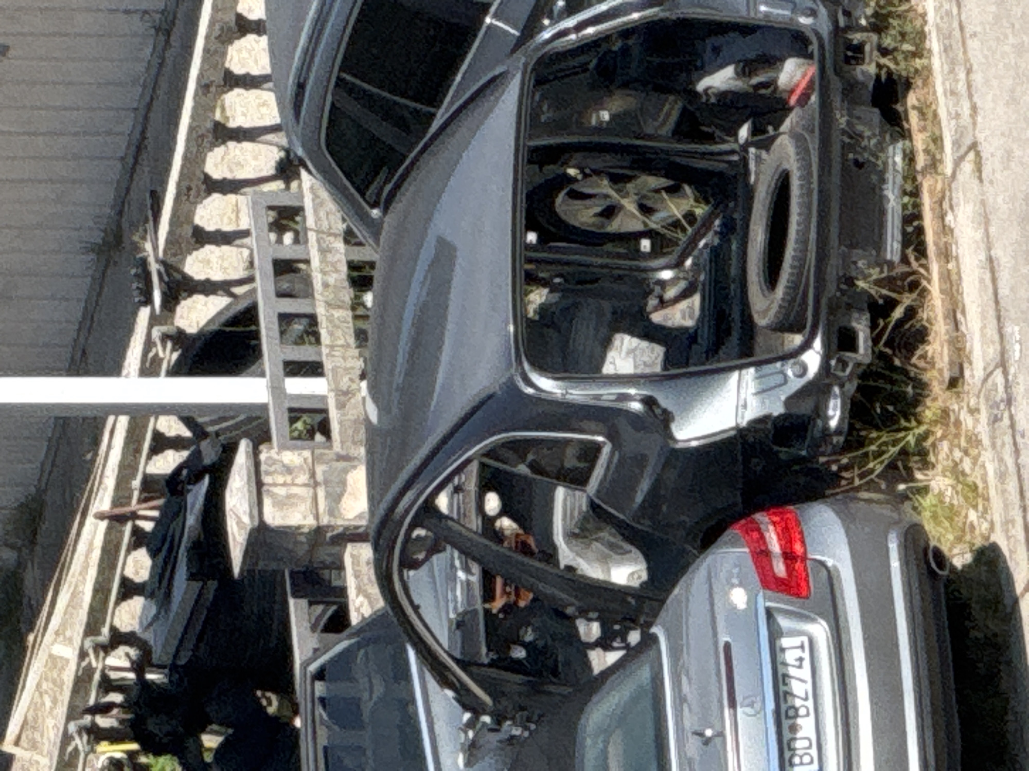 A dark-colored, heavily damaged, four-door sedan, likely a Mercedes-Benz based on the visible license plate "BD BZ741", is parked at an angle in an outdoor area. The vehicle appears to be stripped of many parts, with its doors open and interior exposed. A spare tire is visible in the rear cargo area. The car is surrounded by dry grass and dirt, with a stone wall and fence in the background. Adjacent to the damaged sedan is another vehicle, also appearing to be a dark-colored sedan, partially visible. The scene suggests a junkyard or an area where discarded vehicles are stored. The presence of the "BD" license plate prefix indicates the vehicle is registered in Budva, Montenegro.