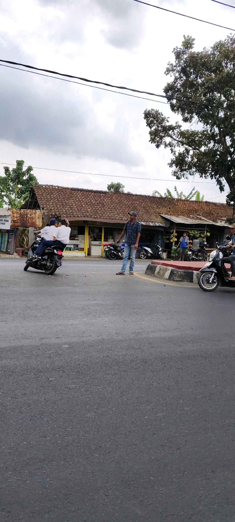 The image shows a street scene in Tasikmalaya, Indonesia, under a cloudy sky. A man wearing a plaid shirt and jeans is standing on the right side of the road, facing left, with his right arm extended. He appears to be directing traffic or interacting with a group of people.
In the center of the image, two individuals on a black motorcycle are turning. They are both wearing white shirts. Behind them, a building with a brown tiled roof and yellow accents is visible, with several motorcycles parked in front of it.
To the far left, a sign with Indonesian text is partially visible. The road is paved, and a curb with red and white markings is present on the right side. A large tree with green foliage is seen in the upper right corner of the frame, with power lines crisscrossing the sky.