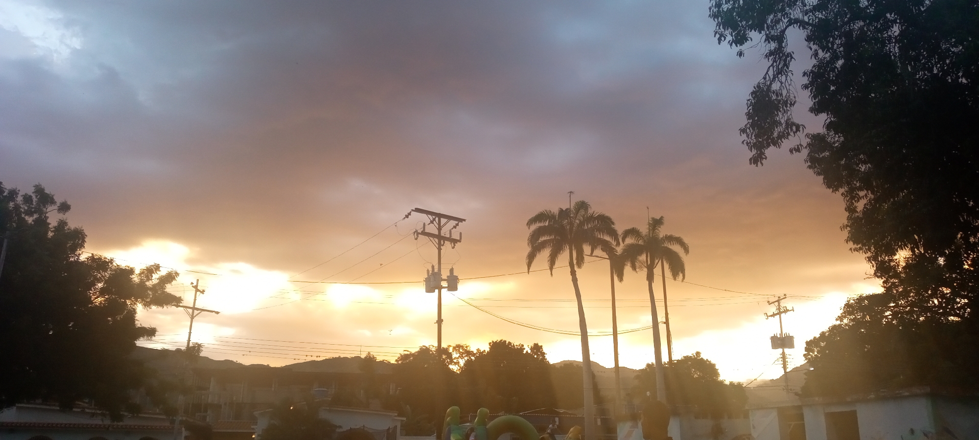 In El Limón, Venezuela, the sky is filled with dramatic, cloud-laden sunset colors ranging from deep grays to vibrant oranges and yellows. Silhouetted against this backdrop are several prominent features: a tall utility pole with transformers and power lines stretching across the frame, and a cluster of three tall palm trees with their fronds clearly defined. In the foreground and to the sides, the dark foliage of trees partially obscures the scene. Below the sky, a low-lying landscape is visible, with the tops of buildings, including what appears to be a structure with an arched facade, peeking through. The overall impression is a twilight scene in a tropical urban or semi-urban environment.