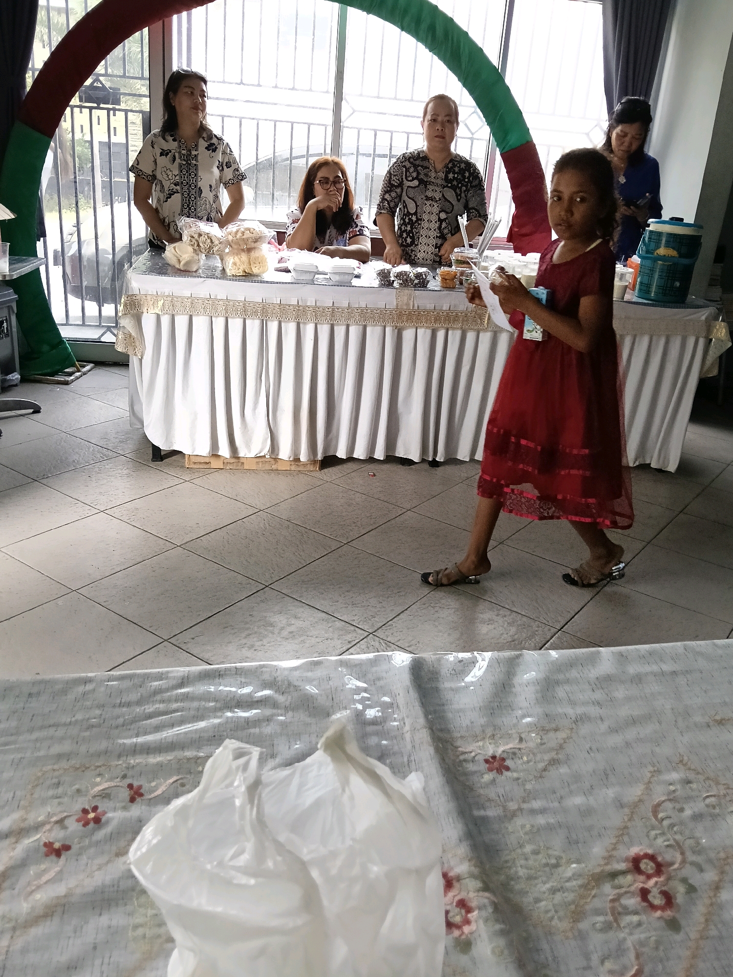 In Jatimulya, Indonesia, an indoor scene depicts a gathering with food items displayed on tables. Four women are visible. Two women stand behind a table covered with a white tablecloth and adorned with lace trim. They appear to be arranging or serving food products. Another woman is seated at the same table, looking towards the side, and seems to be engaged in a conversation. In the foreground, a young girl in a red dress walks past the table, holding a white paper. The room has tiled flooring and large windows in the background, suggesting it could be a community hall or a similar venue. A decorative arch structure with red and green accents is partially visible on the left side of the image.