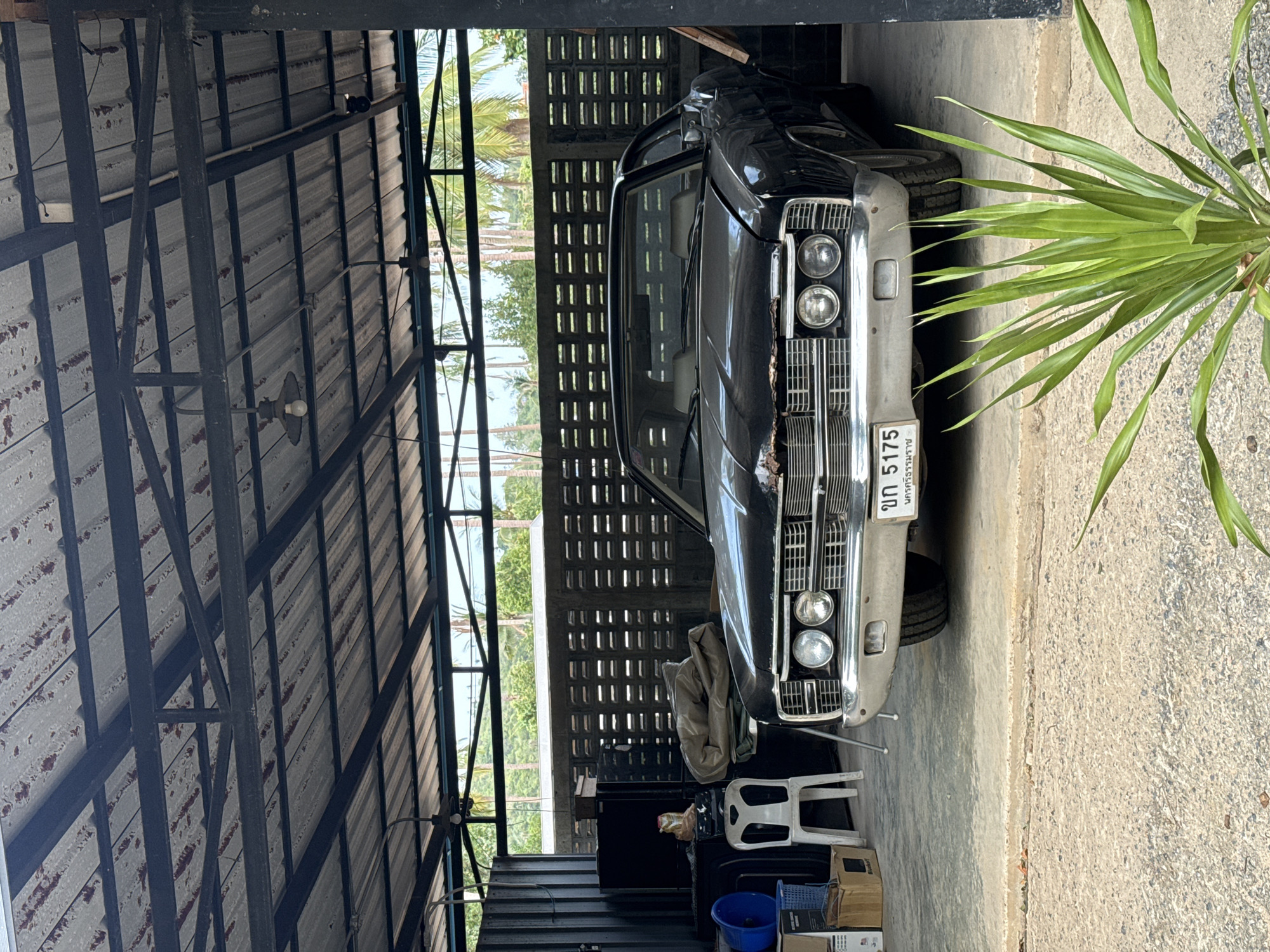 The image depicts a black vintage American car parked under a corrugated metal roof structure. The car is a two-door coupe with chrome detailing and a license plate that reads "ขก 5175". Behind the car, a textured concrete block wall with horizontal openings is visible. To the left of the car, there are stacked boxes, a blue bucket, and a white plastic chair. The overall scene suggests an outdoor parking or storage area, possibly in a more rural or less developed part of Ko Samui District, Thailand, given the tropical foliage visible in the background and the style of the building. There are no people or animals visible in the image. The lighting suggests it is daytime, with the sun casting shadows.