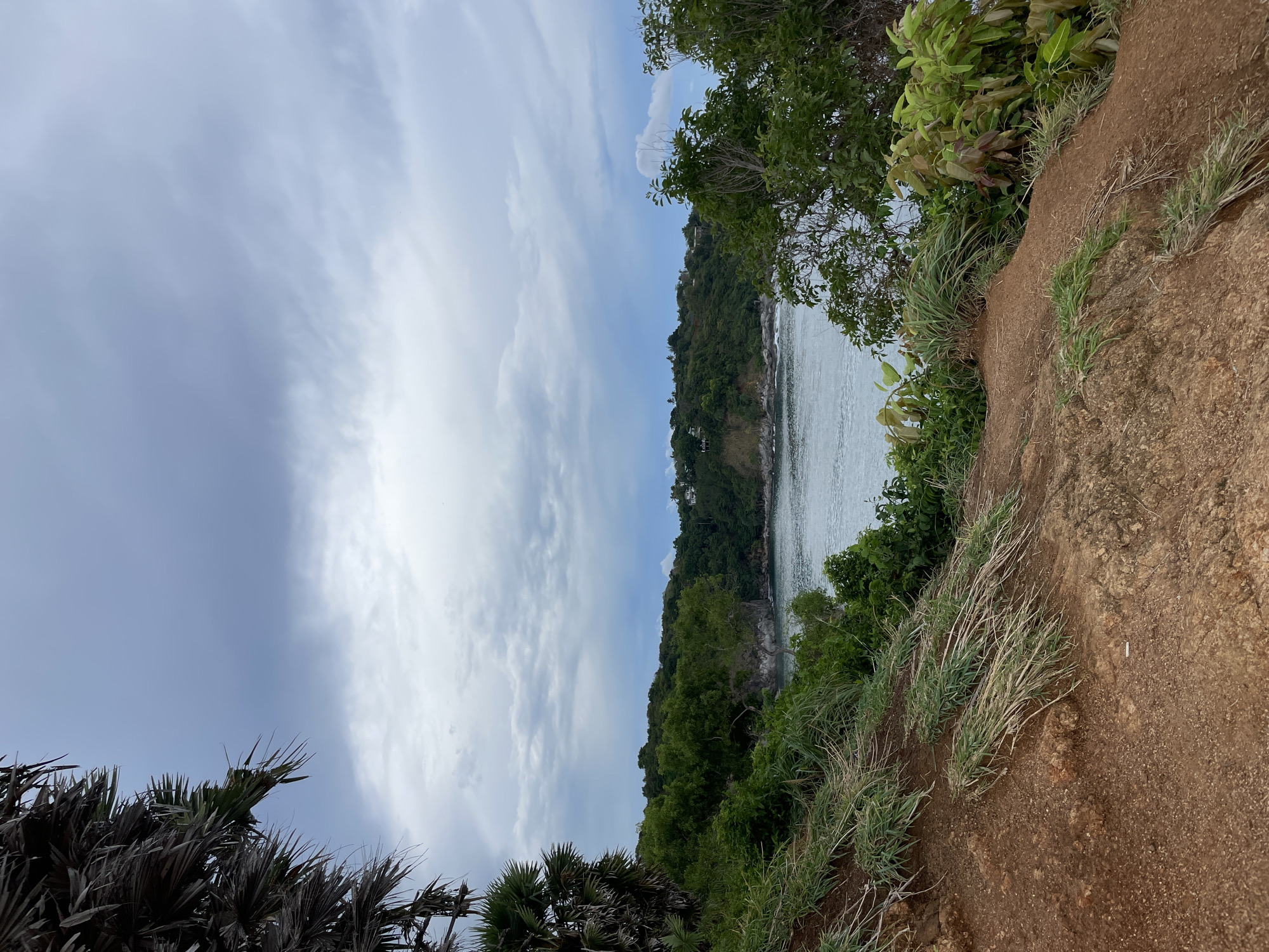 The image depicts a coastal scene in Rawai, Thailand, featuring a view from a high point, likely a cliff or hill. The foreground shows a reddish-brown dirt embankment covered with sparse grass and vegetation. Beyond the embankment, the view extends towards a body of water, possibly the ocean, with land covered in dense green vegetation across the bay. The sky is partly cloudy, with a mix of white and grey clouds dominating the scene. The overall impression is one of a natural, slightly elevated viewpoint overlooking the Rawai coastline.