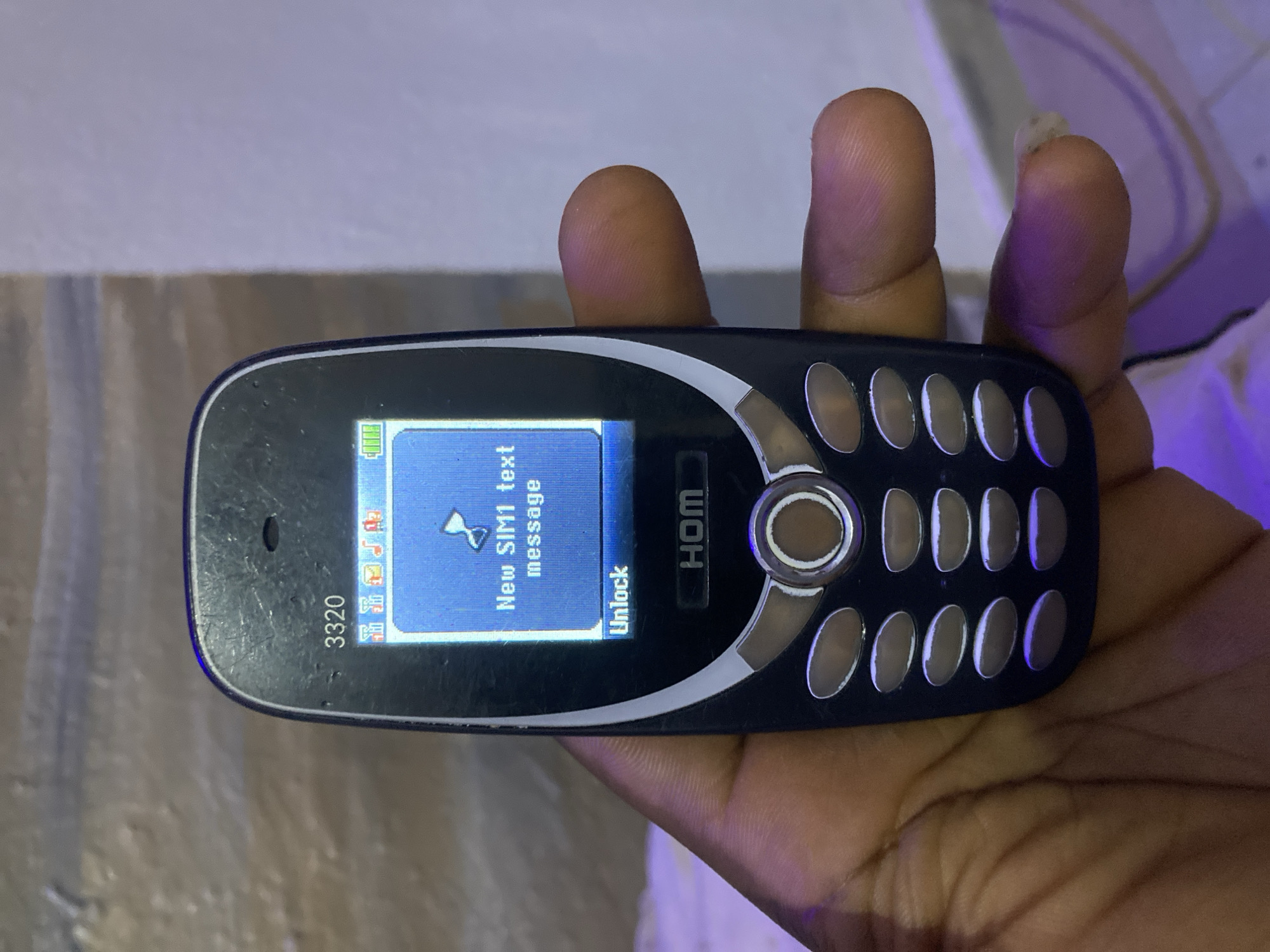 A hand holds a black "HOM 3320" feature phone with a blue screen displaying "New SIM1 text message". The phone has a circular navigation button and a keypad with oval buttons. The phone indicates full battery. The surrounding environment is indistinct, preventing any conclusion about Jos, Nigeria.
