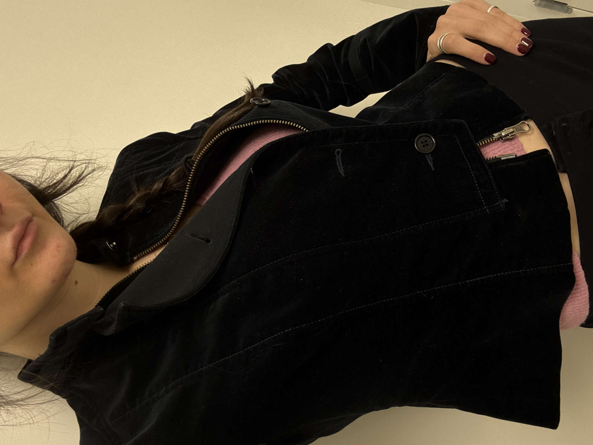 The image displays a close-up, waist-up view of an individual positioned against a plain, light-colored wall. No specific location within the city of New York, United States of America, is identifiable from the visual information presented. The subject is wearing a black velvet jacket, unbuttoned and partially unzipped from the top, revealing a pink ribbed top underneath. The jacket features a stand-up collar and three unfastened black buttons on the visible placket. The individual's left hand, adorned with dark red nail polish and two silver rings (one on the index finger, one on the ring finger), rests on dark trousers. A portion of the subject's face, including the lower lip and chin, is visible at the top, along with a mole on the neck and dark hair styled in a braid over the left shoulder. The posture suggests the individual is posing for a photograph.