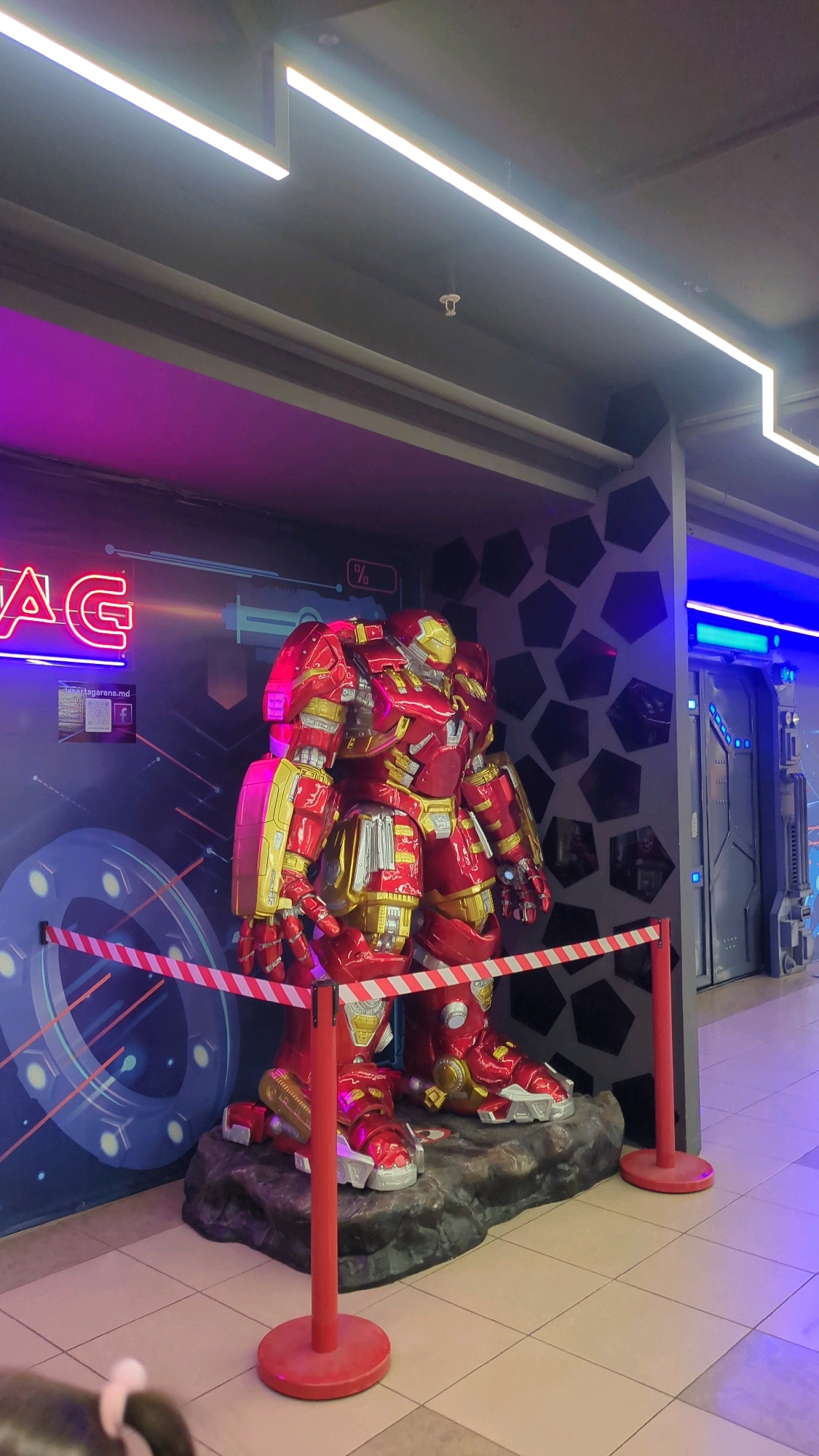 An impressive, life-sized Hulkbuster armor statue stands as the central focal point of this indoor scene. The statue is predominantly red with gold accents, detailed with silver metallic elements, and is positioned on a rocky base. A red and white striped ribbon barrier with red stanchions is placed in front of the statue, suggesting it is an exhibit or a photo opportunity.
Behind the statue, a large wall features neon lights spelling out "LASERTAG" in bright pink letters, accompanied by graphics that appear to be part of a game or promotional material. Further back, a gray wall is decorated with a pattern of dark, irregular hexagons. To the right of the Hulkbuster, a futuristic-looking doorway or entrance is illuminated with blue LED lights, suggesting a distinct area within the establishment. The floor is tiled in a light beige color, and overhead, modern linear fluorescent lights are visible, with a fire sprinkler head also mounted on the ceiling.
The overall environment suggests an entertainment venue, likely an arcade or laser tag arena, given the prominent "LASERTAG" signage. Based on the website URL displayed on a small sign to the left of the "LASERTAG" sign, which reads "lasertagaerna.md," this location is confirmed to be in Chișinău, Moldova. The presence of the statue implies a thematic decoration designed to attract visitors, possibly to an area associated with comic book or superhero franchises. There are no visible people in the frame.