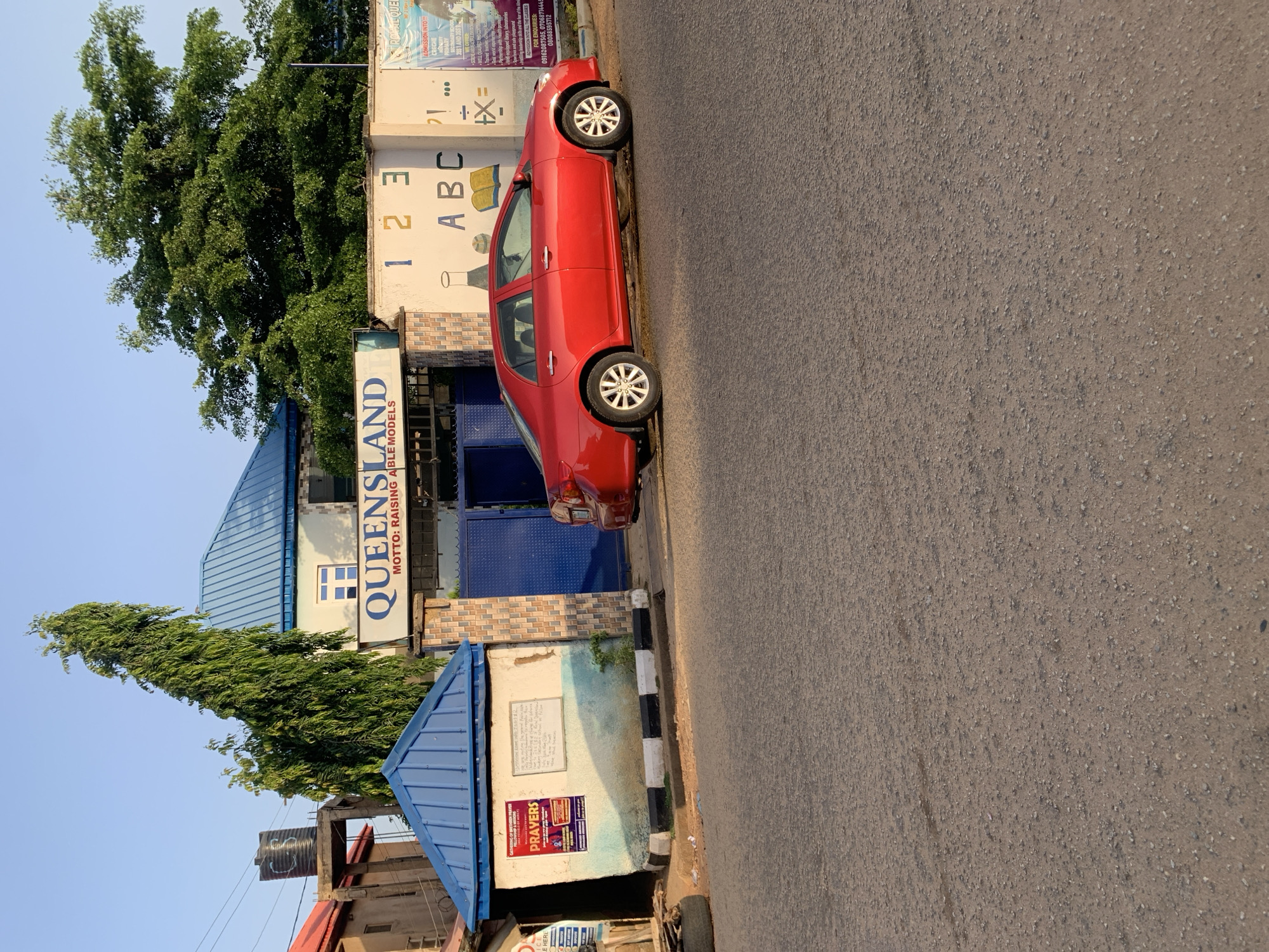A red car is parked on the side of a road in front of the QueensLand School in New Stadium, Nigeria. The school is a light-colored building with a blue gate and a sign that reads "QUEENSLAND MOTTO: RAISING ABLE MODELS". The image shows some trees near the school building. The sky is visible at the top of the image. The street is an asphalt street. A water tank is visible in the background.