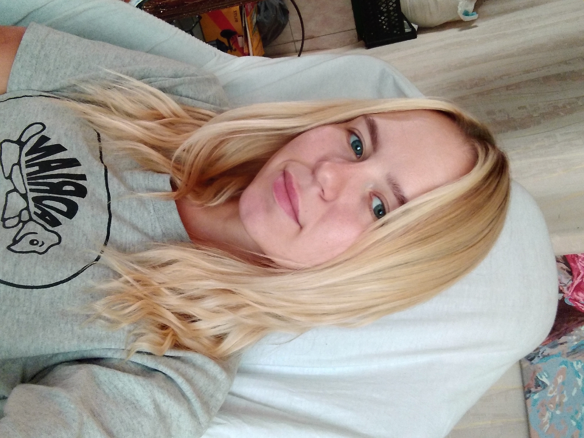 A young woman with blonde hair and blue eyes is sitting in a chair, wearing a grey t-shirt with a graphic design. She is looking directly at the camera, with her head slightly tilted. The background appears to be indoors, with a beige curtain and a white blanket draped over the chair. There are some boxes and a black crate visible in the background, suggesting a cluttered or storage area. The image was taken in Radisne, Ukraine.