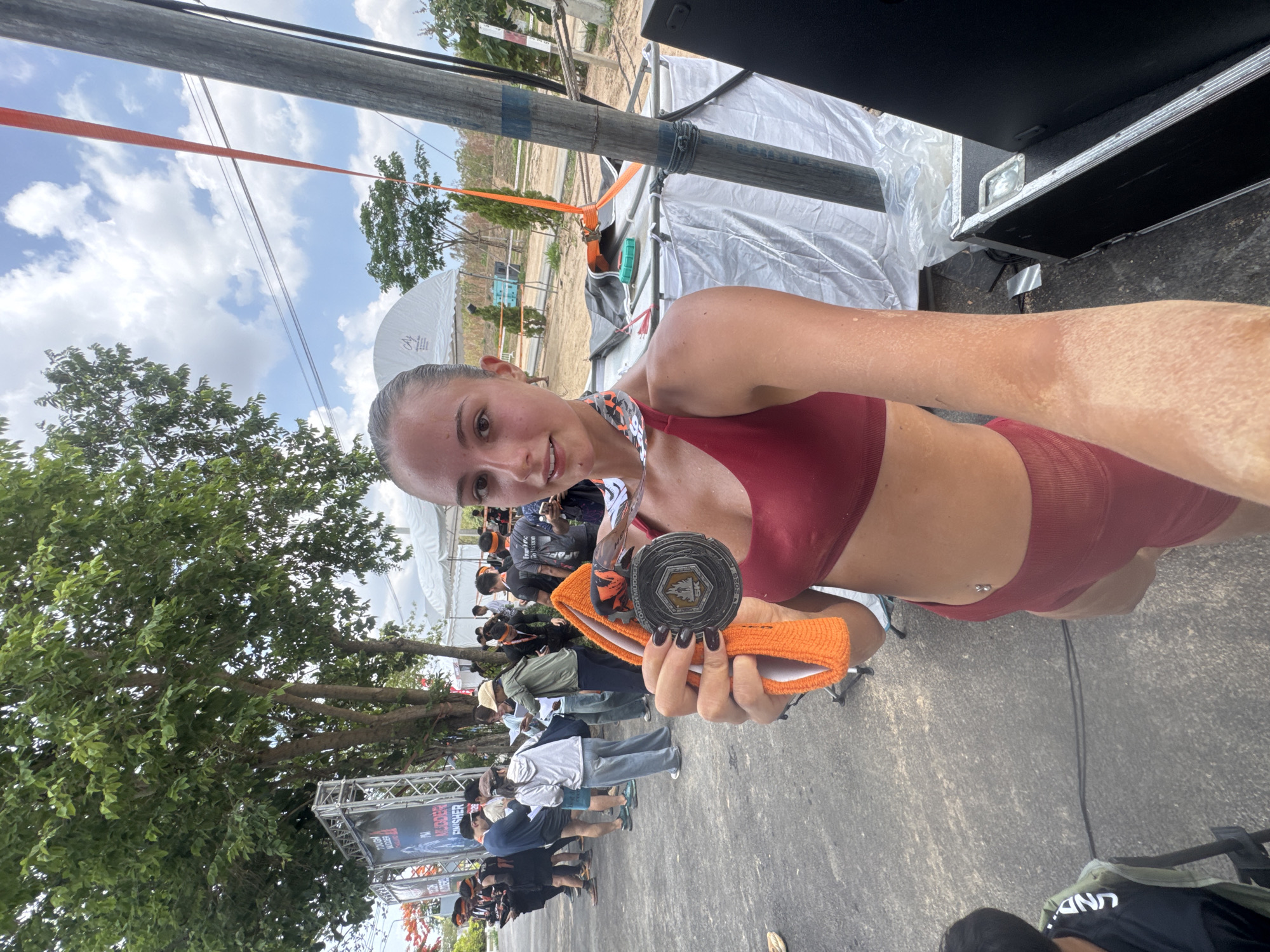 A woman is holding a medal in her right hand, looking into the camera and smiling. She has a maroon sports bra and shorts and the medal is silver with an orange band. Behind her, other people are standing in line. There is a tree and a banner that reads "Mudder Finisher". There is a white tent in the background. The location is บ้านสำนักตะแบก, Thailand.