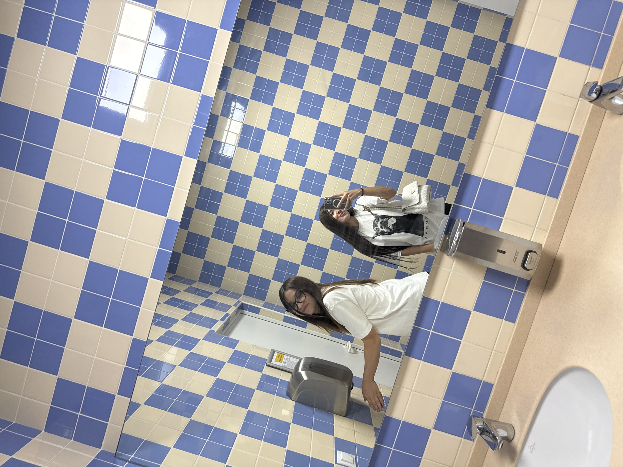 A young woman with long dark hair and glasses is reflected in a mirror in a bathroom. She is wearing a white t-shirt and is reaching out her left hand towards a silver automatic hand dryer mounted on a blue and beige checkered tiled wall. To her right, a second reflection of the woman is visible, this time without glasses, wearing a white t-shirt with a graphic print and a white purse. She is holding a smartphone and taking a selfie. The bathroom environment features a sink with a chrome faucet, a soap dispenser, and a checkered tile pattern on the walls and a section of the floor. The location is identified as Bardar, Moldova.