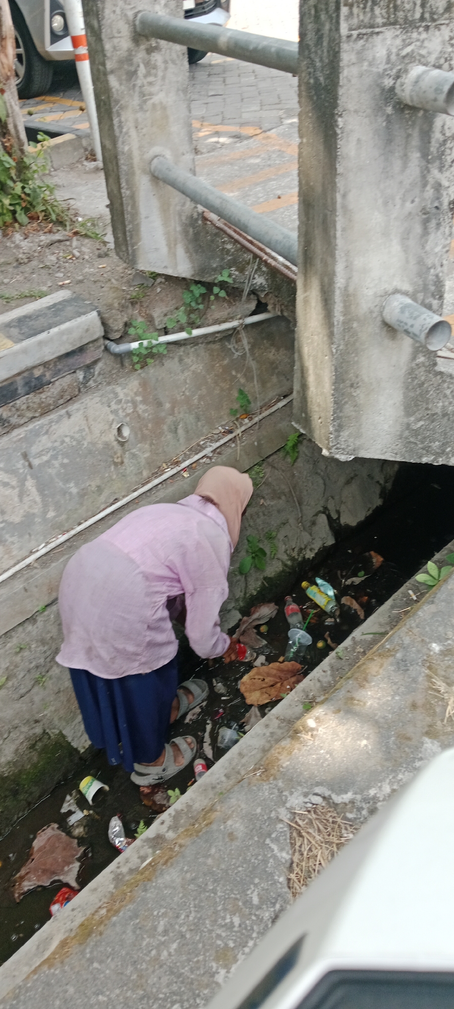 In Maguwoharjo, Indonesia, a person wearing a pink long-sleeved shirt and a blue skirt, with a beige hijab, is crouched down and reaching into a dirty drainage ditch. The ditch contains a significant amount of trash, including plastic bottles, wrappers, leaves, and other debris. The person appears to be sifting through the trash. The surroundings are urban, with concrete structures, a paved surface with yellow markings, and some vegetation growing in and around the ditch. A portion of a white vehicle is visible in the foreground.