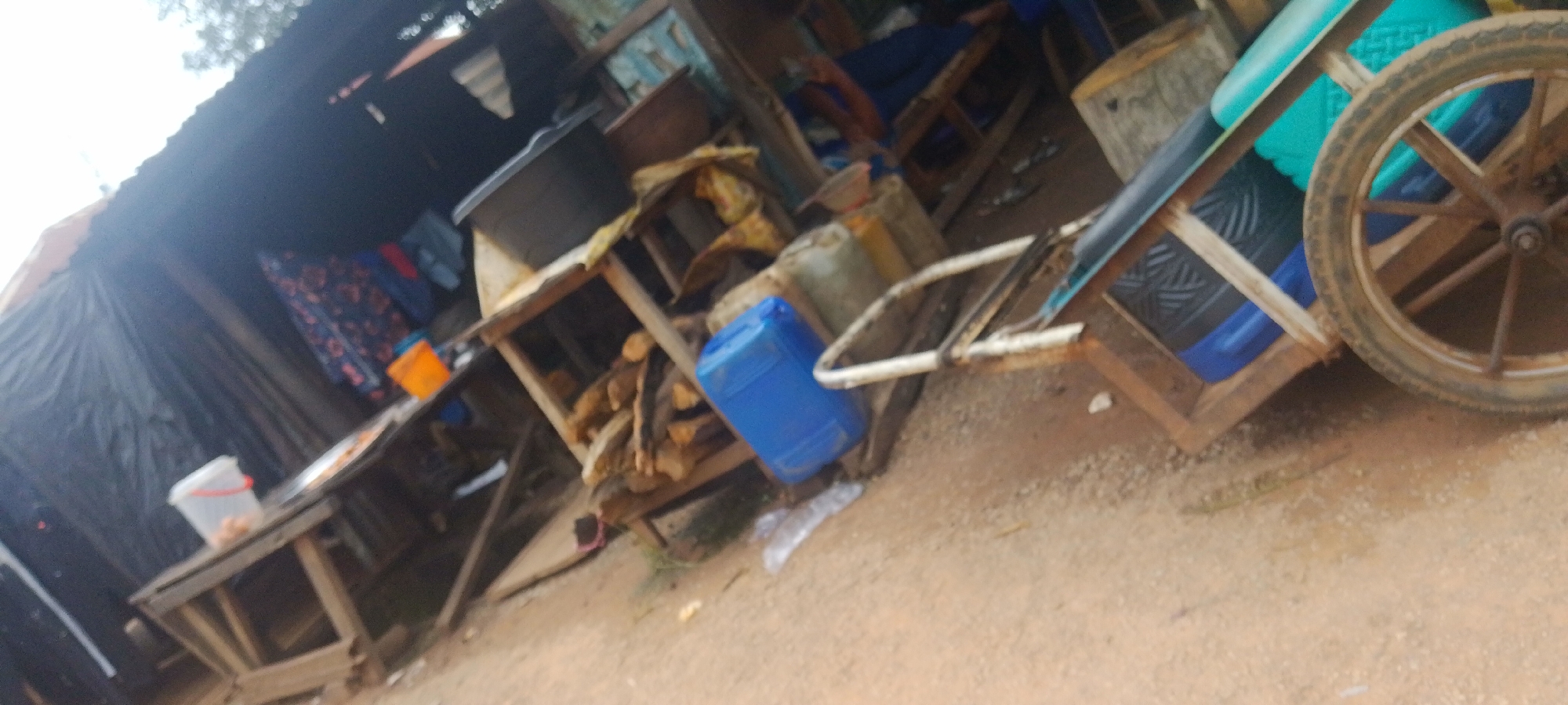 The image depicts an outdoor market stall in Baissa, Nigeria. The scene is cluttered with various objects, suggesting a busy commercial setting. A wooden cart with a spoked wheel is prominently featured, loaded with blue and teal containers and a black item with a patterned surface. To the left of the cart, a wooden table holds a white bucket containing small, light-colored items, possibly food. Behind this table, a black tarp covers part of a stall, and what appears to be patterned fabric is visible. Further to the left, a rustic, open-air shelter is present, with corrugated metal roofing and wooden supports.
Another wooden structure, possibly a stall or shelving unit, holds stacks of firewood and a large blue plastic container. In the background, other indistinct structures and items are visible, contributing to the impression of a vibrant market. The ground is sandy or dusty. The lighting suggests it is daytime, and there are no clear indicators of specific weather conditions or emotions. No visible text is present in the image. The overall impression is that of a functional, perhaps makeshift, market setup for selling goods.