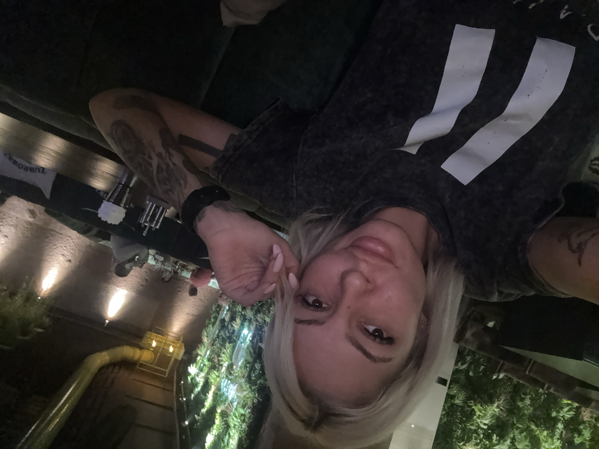 A woman with blonde hair and tattoos on her arms is taking a selfie. She is wearing a dark grey shirt with two white stripes. She is at a restaurant. Outside the restaurant is greenery, and a yellow pipe. The photo was taken in the city of Plovdiv, Bulgaria.