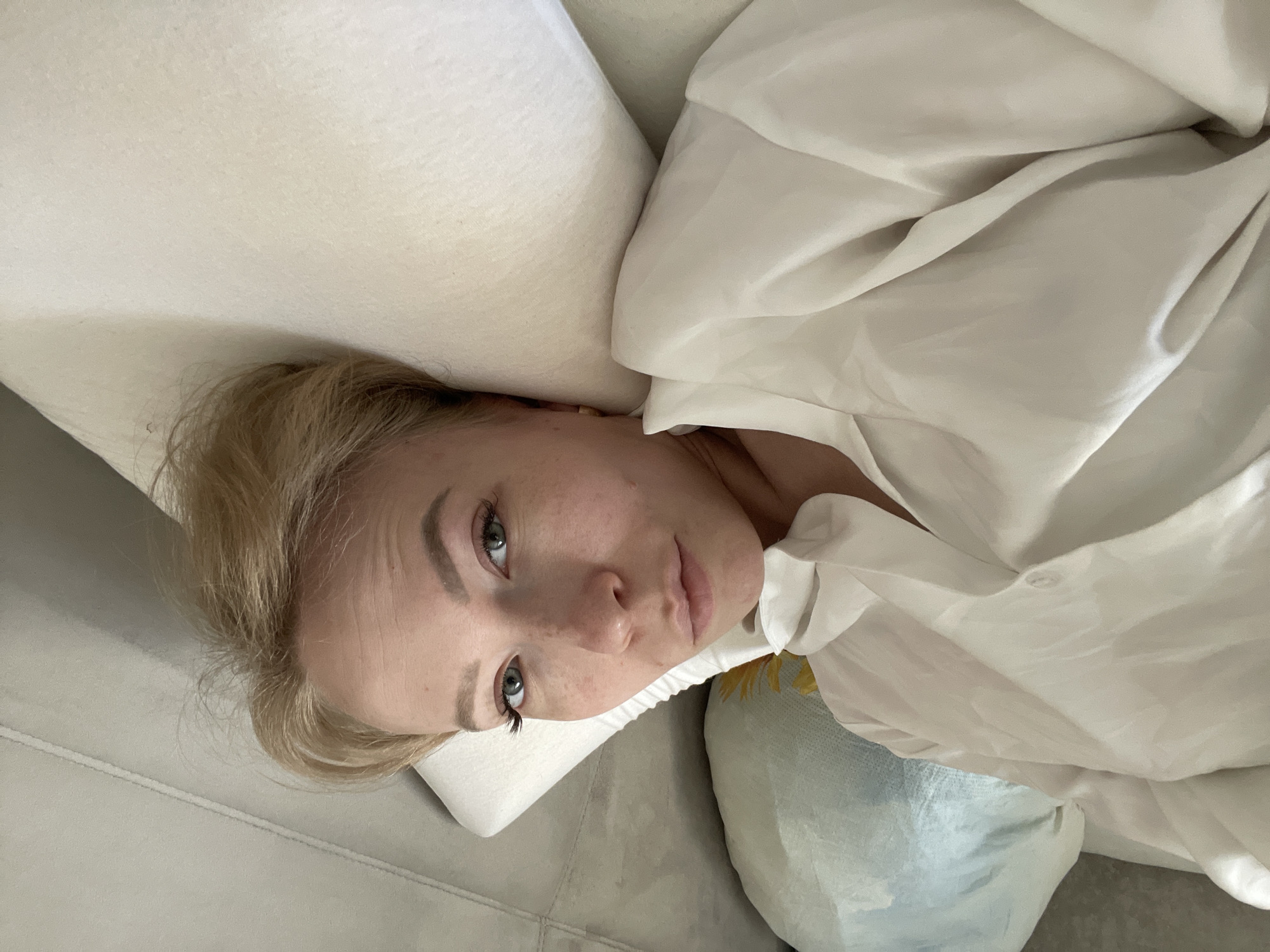 A young, fair-skinned woman with blonde hair is lying down, possibly on a sofa or bed. Her head rests on a white pillow, and she is partially covered with a white, wrinkled shirt or sheet. The surface she's lying on appears to be a light gray or beige color. The background and surrounding environment are not clearly visible, but the scene appears to be indoors. There is nothing to suggest the image was taken in the city of Perm, Russia.