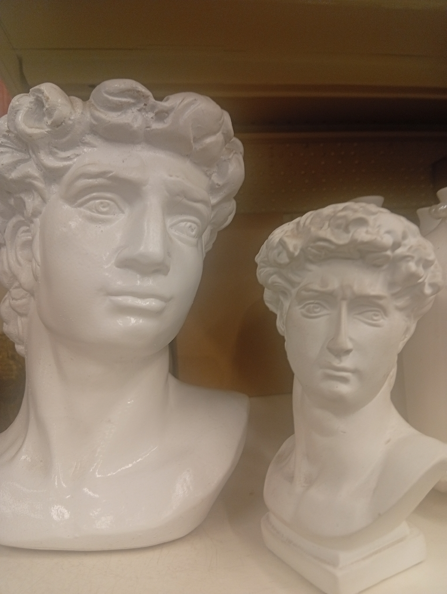 Two white plaster busts of men are displayed on a shelf. The bust on the left is larger and more detailed, featuring curly hair, eyes, a nose, and lips. The bust on the right is smaller and slightly more abstract, with less defined features. The background is a blurred wooden surface. The image is an indoor shot with soft lighting. This scene is from an unknown location in the city of Pushkino, Russia.