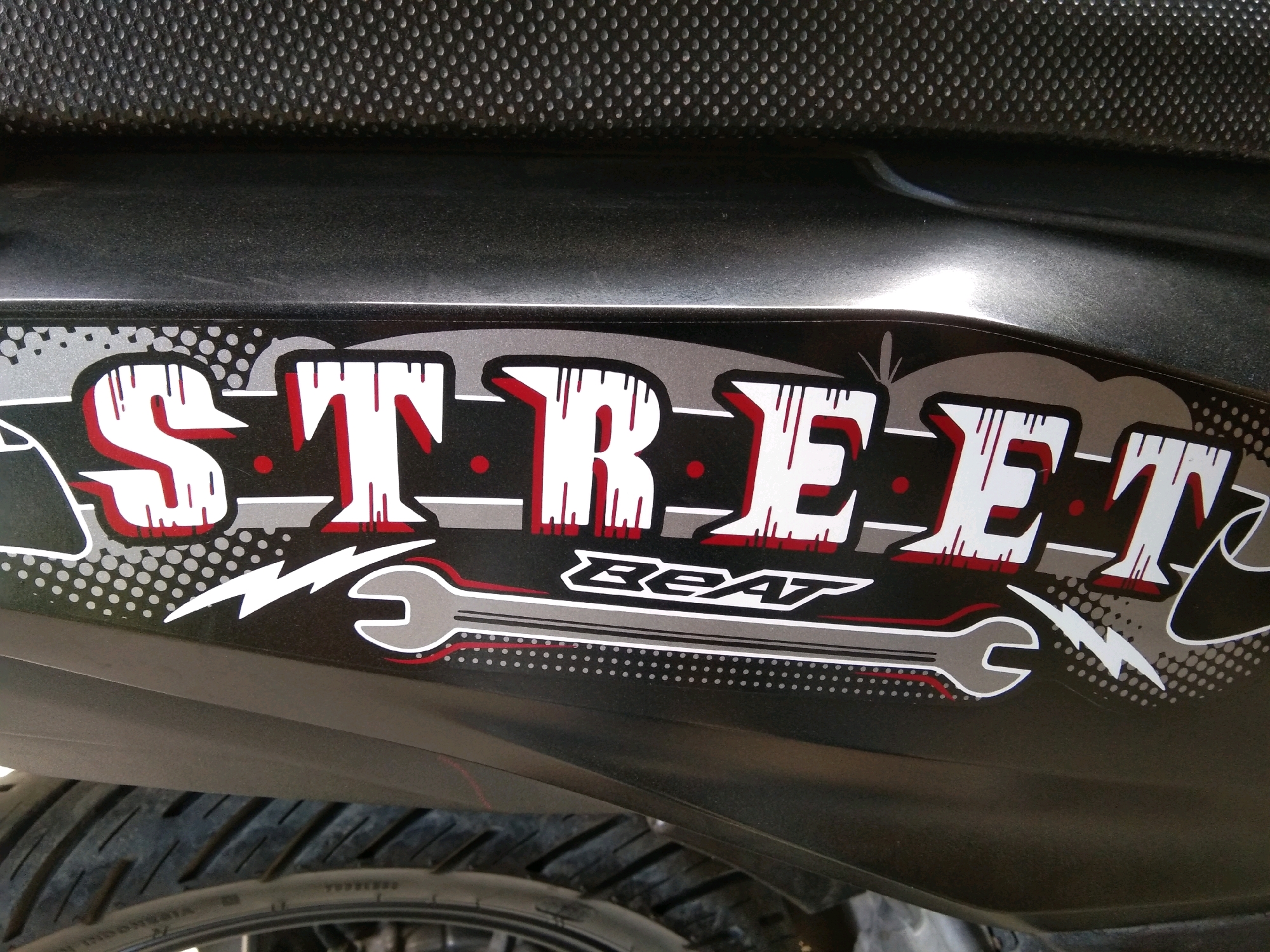 The image shows a close-up of a black motorcycle's side panel with a decal. The decal features the word "STREET" in large, stylized white letters with red outlines and black shading. Below "STREET", the word "BEAT" is written in a smaller, more angular font, also with white lettering and red and grey accents. The decal is set against a black background and incorporates graphic elements such as silver dots, red circles, lightning bolts, and wrench-like shapes. The texture of the motorcycle's seat is visible at the top of the frame, and a portion of the rear tire is visible at the bottom. The scene is an outdoor setting. The image is taken in the city of Karangsambung, Indonesia.