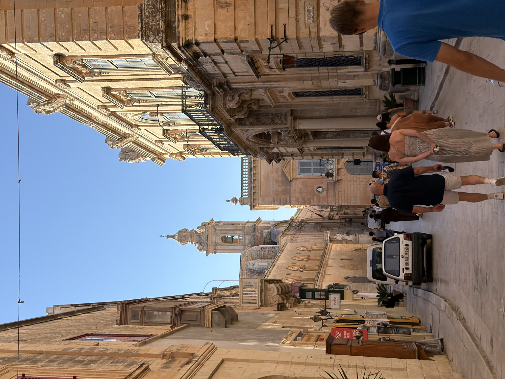 The image depicts a street scene in Mdina, Malta. The architecture is characterized by old, sun-drenched stone buildings with ornate details and balconies. A clear blue sky is visible above.
Several people are walking along the street, some in casual attire. In the foreground, a man in a blue t-shirt is partially visible. Further down the street, a group of people are walking together. A white car is parked on the side of the road, with a license plate visible.
The scene appears to be set during the daytime, with bright sunlight casting shadows. The atmosphere seems relaxed, suggesting a tourist or leisurely walk through the historic city. The buildings suggest a Mediterranean or European historical setting.
Visible text includes "Malta" on a sign above a shop entrance and a license plate on the car, which reads "BHAA 994". There is also a red sign with text and an image that looks like an ice cream advertisement.