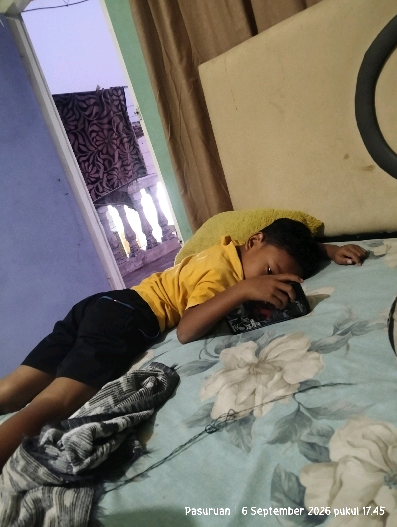 A young boy is lying on a bed covered with a light blue sheet featuring large white floral patterns. He is wearing a yellow t-shirt and black shorts, and is holding a smartphone in his hands, resting his head on a yellow pillow. To the left of the boy, a doorway reveals a glimpse of an outdoor balcony with ornate railings and a patterned fabric hanging. The room appears to be dimly lit, possibly at dusk, with a curtain of a brownish hue visible in the background. A folded grey and white patterned blanket or scarf lies on the bed near the boy's legs. Text in the bottom right corner reads "Pasuruan | 6 September 2026 pukul 17.45", indicating the location as Pasuruan, Indonesia, and the date and time.