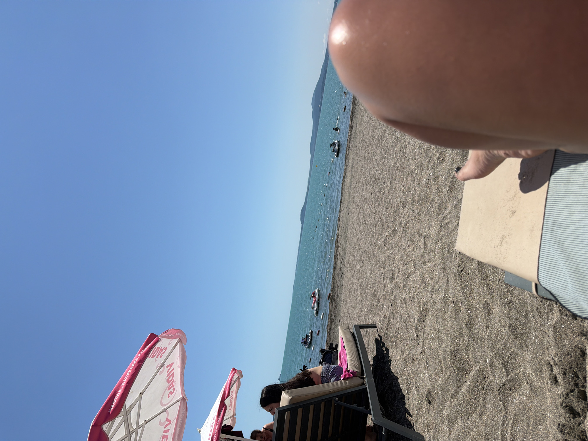 The image depicts a sunny day at a beach in Burgas, Bulgaria. In the foreground, a person's legs are visible, resting on a striped lounge chair. The beach sand is dark and pebbly. Further back, a beach umbrella with pink and white stripes is partially visible, offering shade to a woman sitting on a lounge chair. She is wearing denim shorts and a bikini top, with a pink towel draped over the chair. In the turquoise water, several jet skis are present, and some people are visible in the sea. The horizon is marked by a line of hills under a clear blue sky.