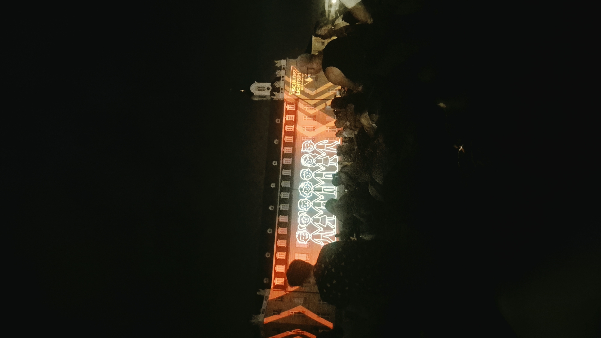 In Karlsruhe, Germany, a nighttime event features a projection on the façade of a historic building, likely a palace or significant public structure. The projection displays a row of glowing, stylized figures of children holding hands, outlined in white light. Orange and yellow geometric patterns are also projected onto the building's exterior, along with text reading "SCHLOSS" and "LICHTSPIEL." A crowd of people is gathered in front of the building, silhouetted against the illuminated projection. The scene captures a moment of public entertainment or a cultural festival in the city.